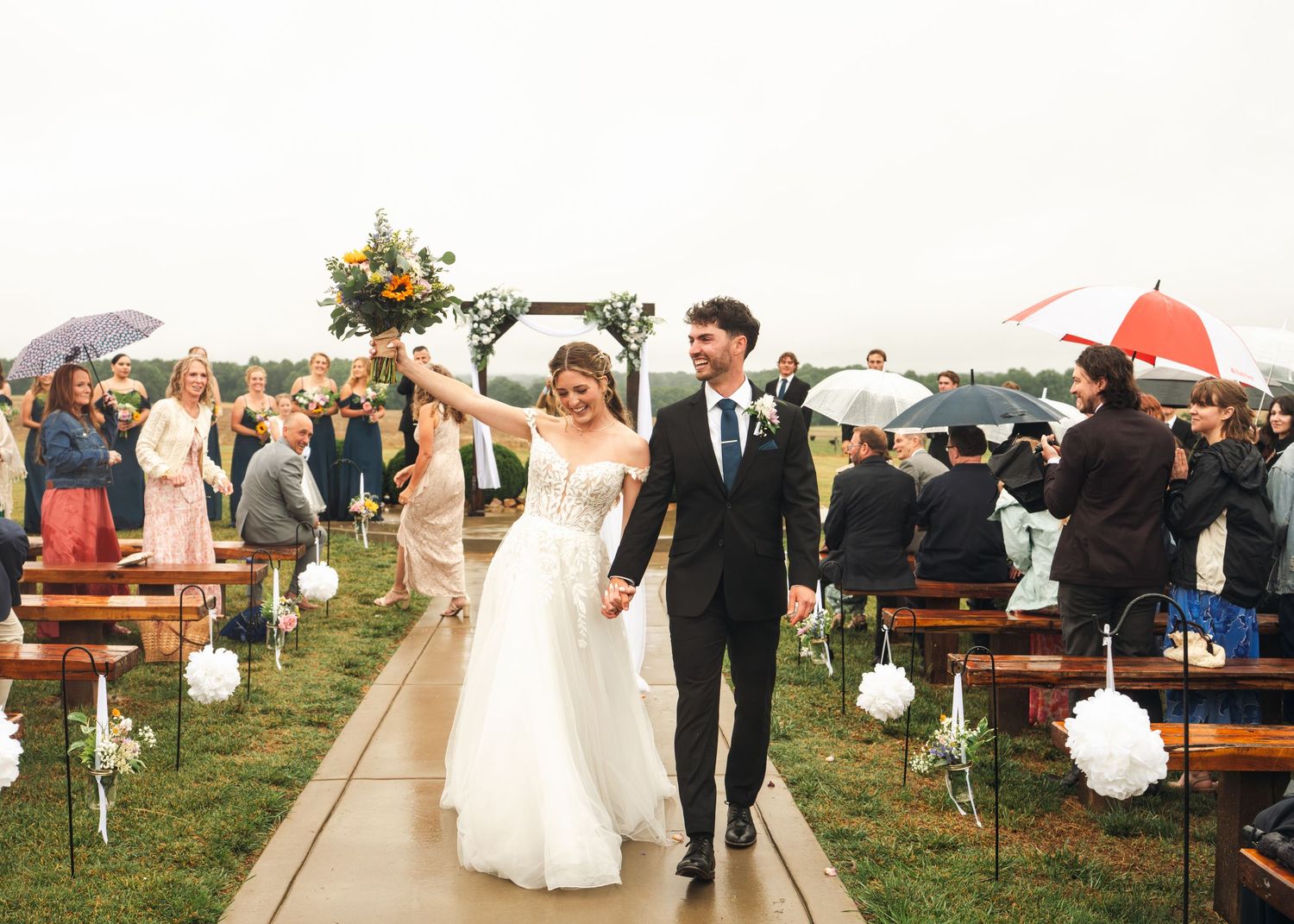Newlyweds walk joyfully down the aisle as guests celebrate with umbrellas at an outdoor rainy wedding.