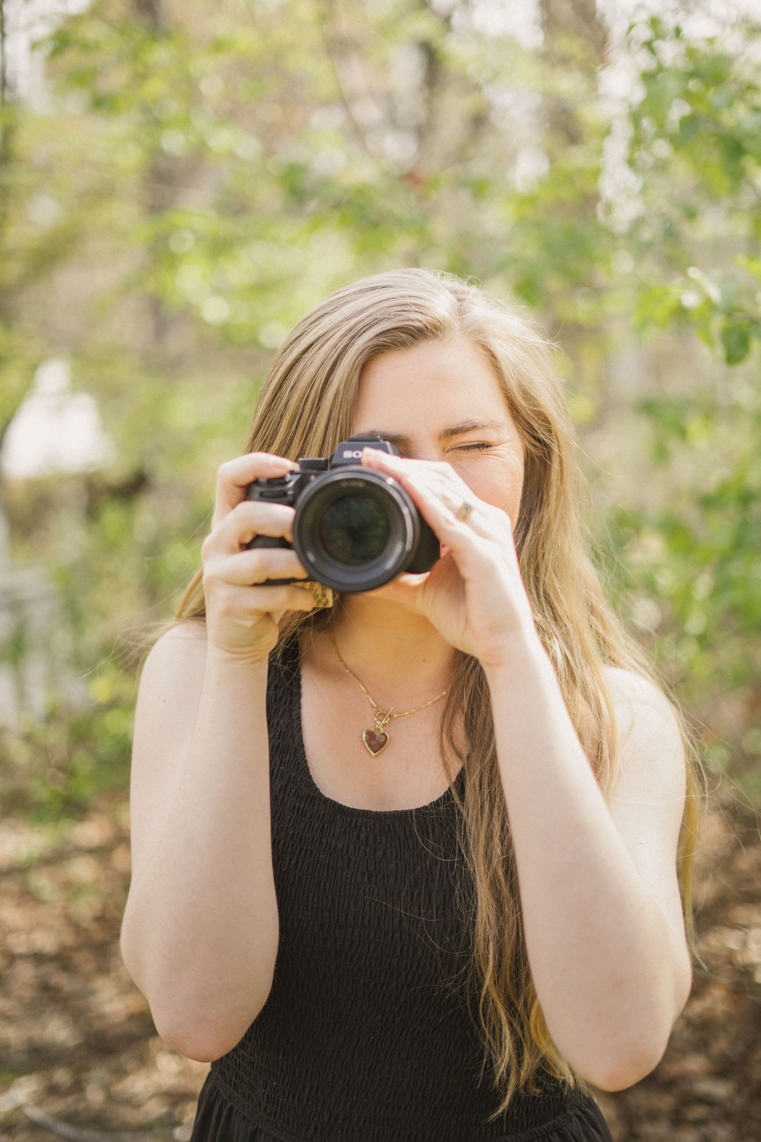 Blonde woman in black dress aiming DSLR camera toward viewer in bright outdoor woodland setting.