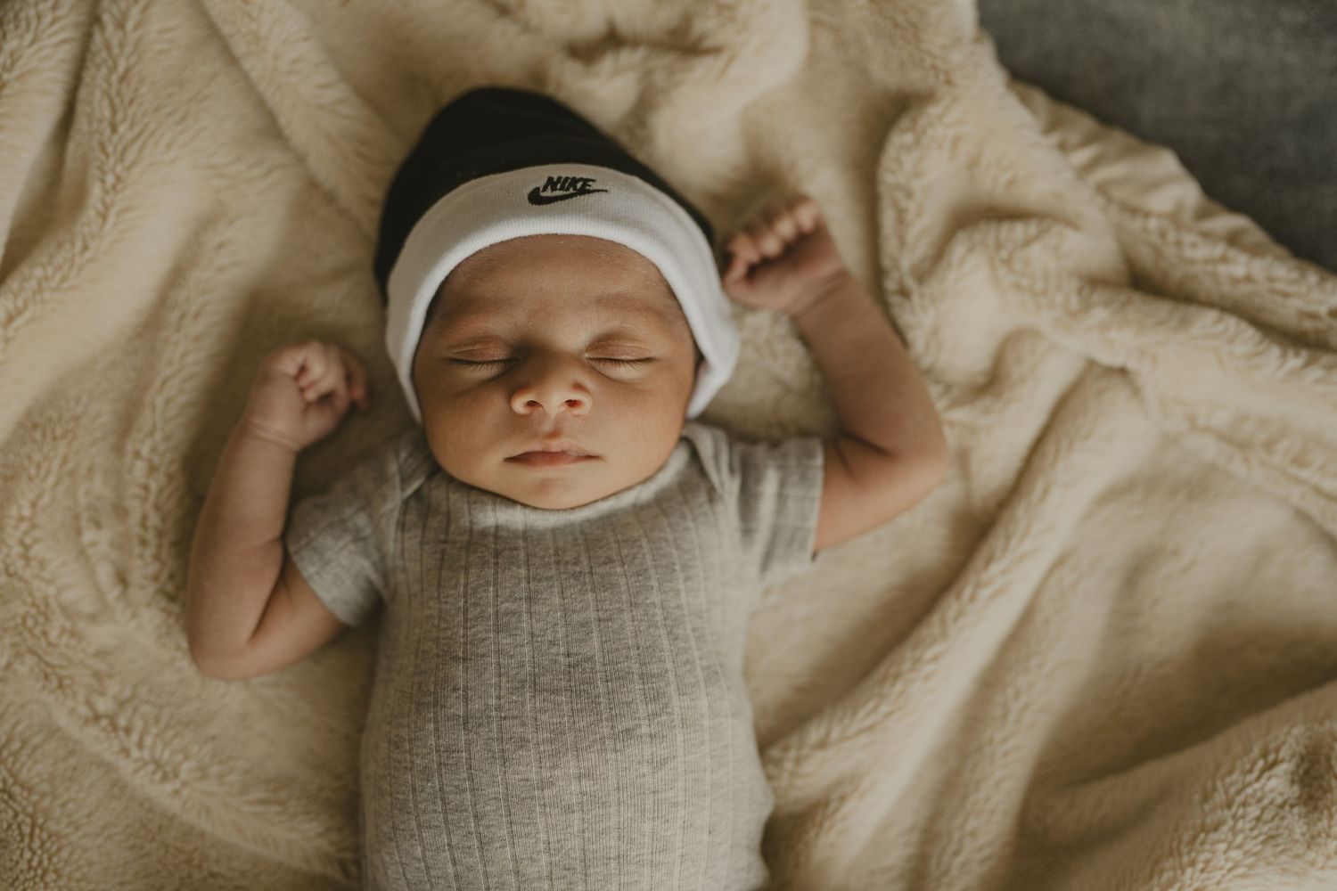 Peaceful newborn baby sleeping in a knit wrap wearing a Nike hat on a cream blanket.