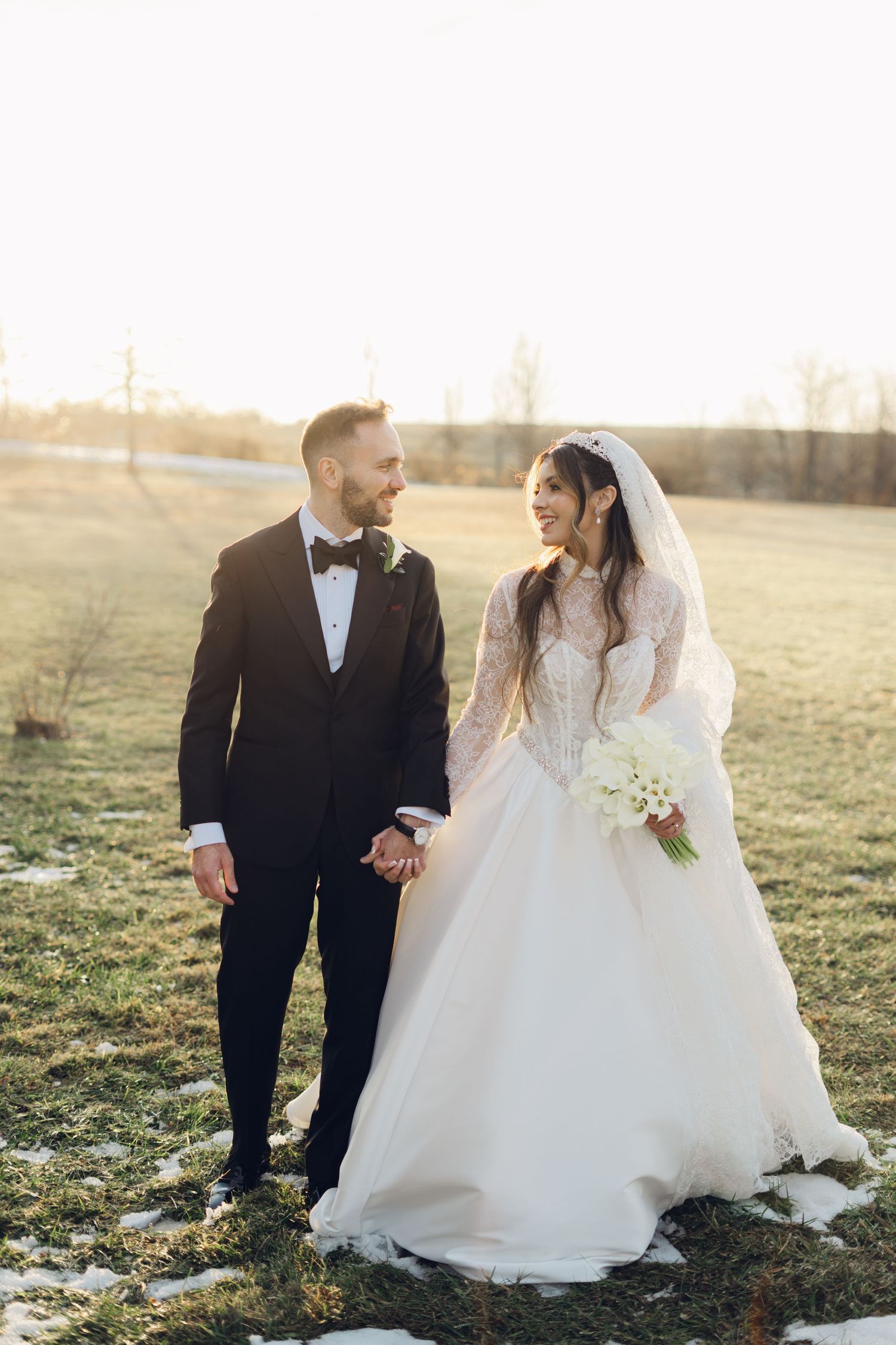 Happy bride and groom walk hand in hand across green field holding white calla lily bouquet at sunset.