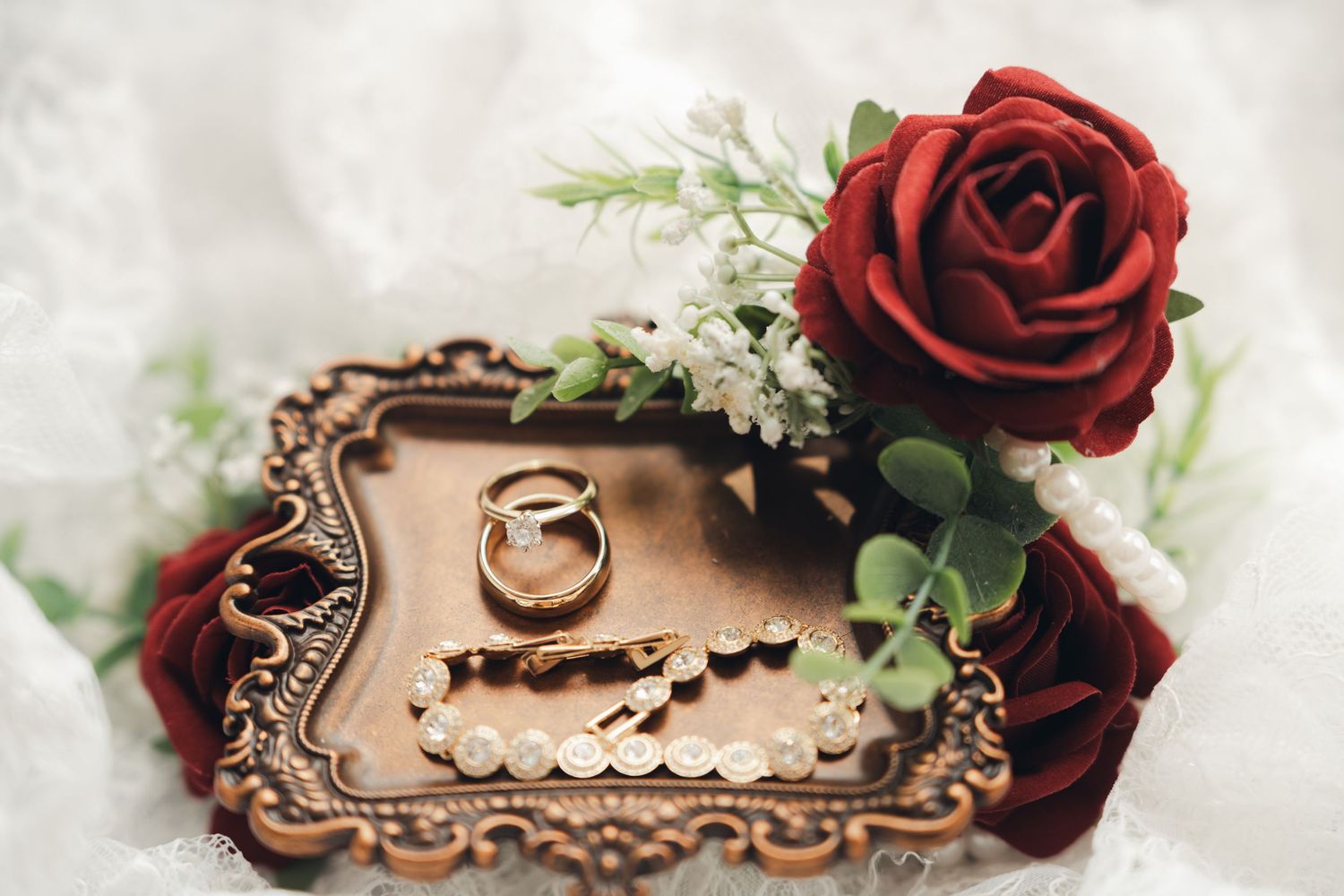 Wedding rings and pearl bracelet displayed on ornate vintage tray with red roses and white flowers.