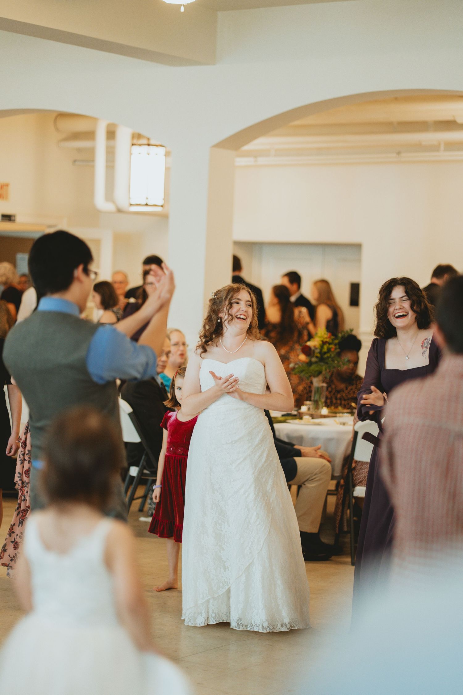 Radiant bride in white lace gown laughs joyfully while dancing with guests at her wedding reception.