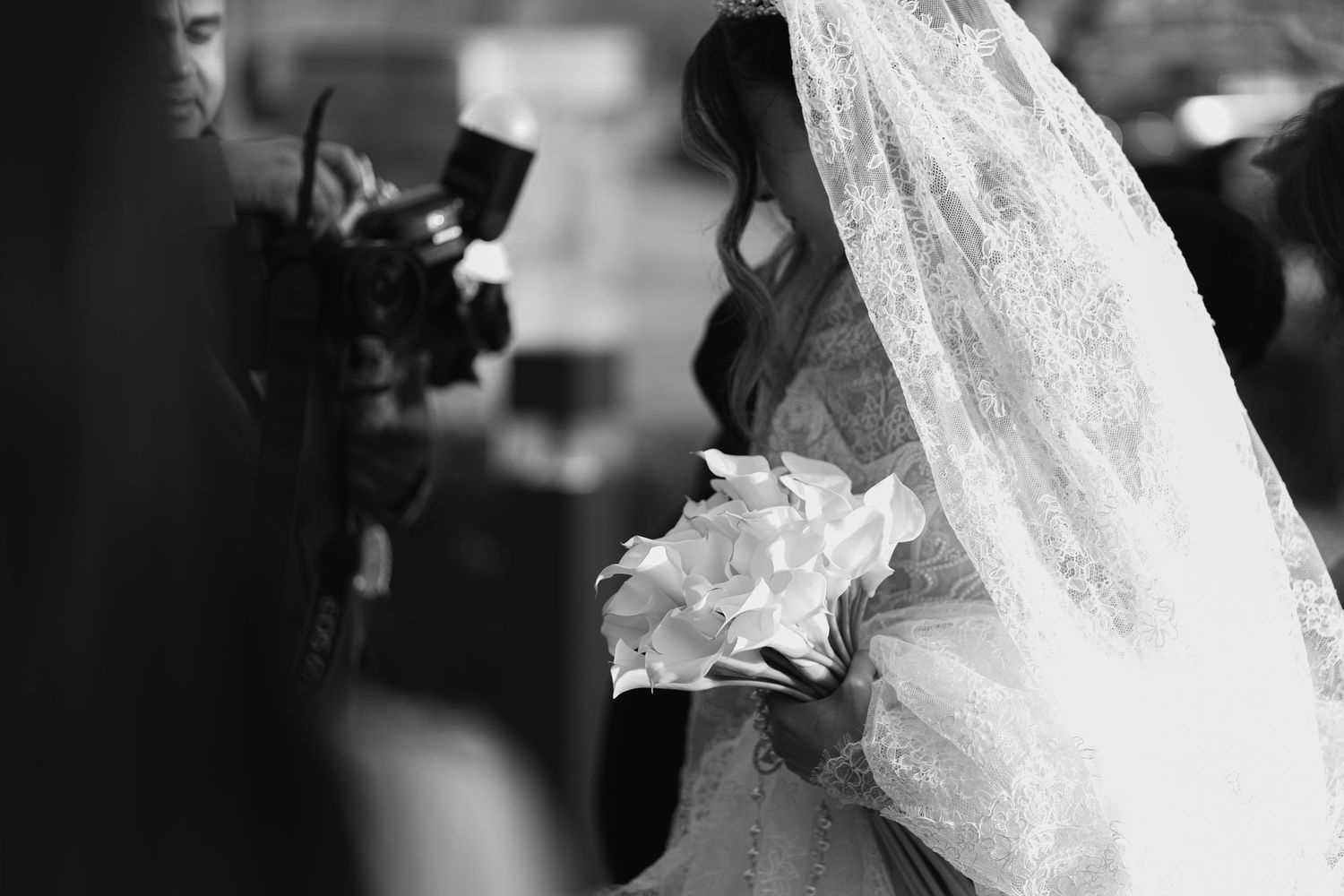 Black and white artistic photo of bride in veil holding white calla lily bouquet at wedding.