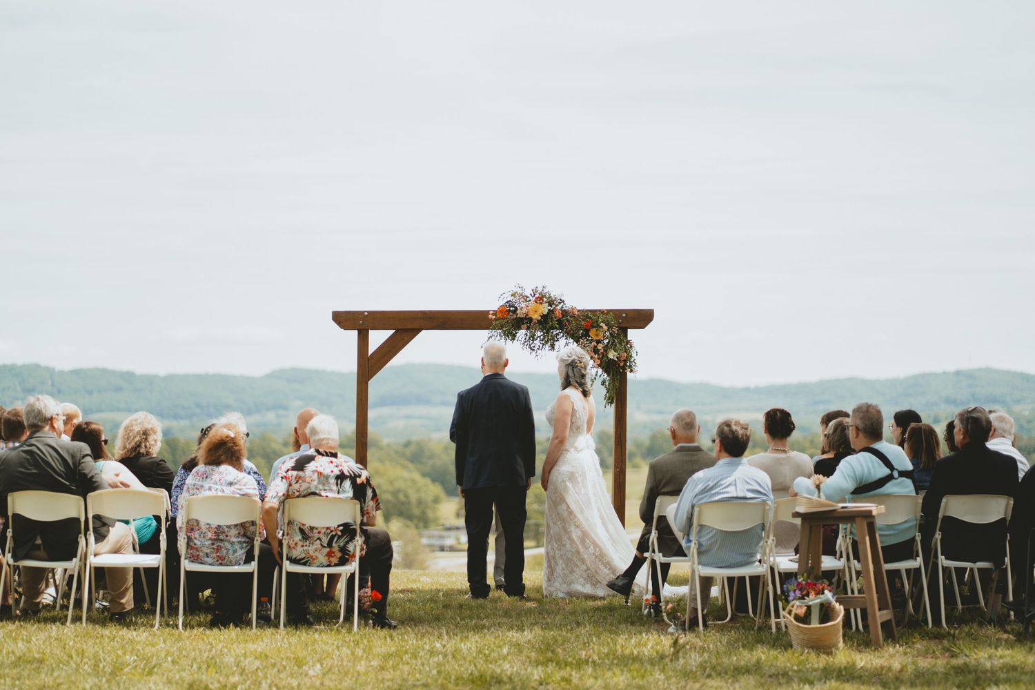 Outdoor wedding ceremony with guests seated facing couple at a wooden arch with floral arrangements.