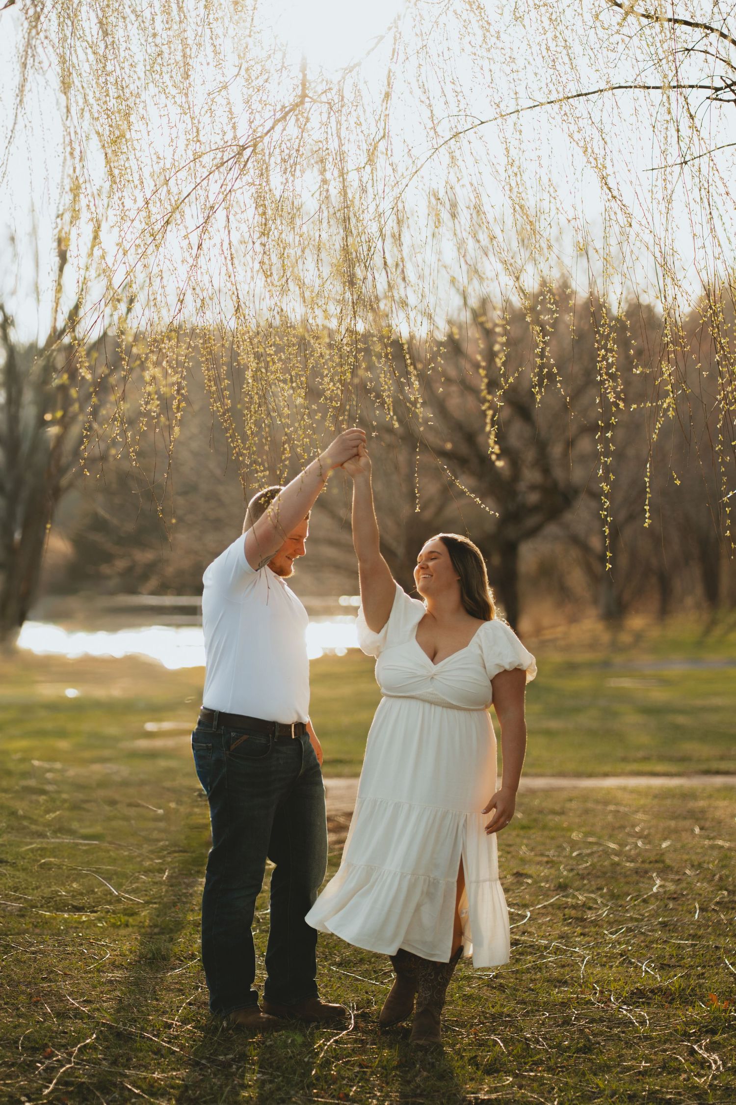 Couple dancing under a weeping willow tree in a sunlit park during golden hour engagement session.