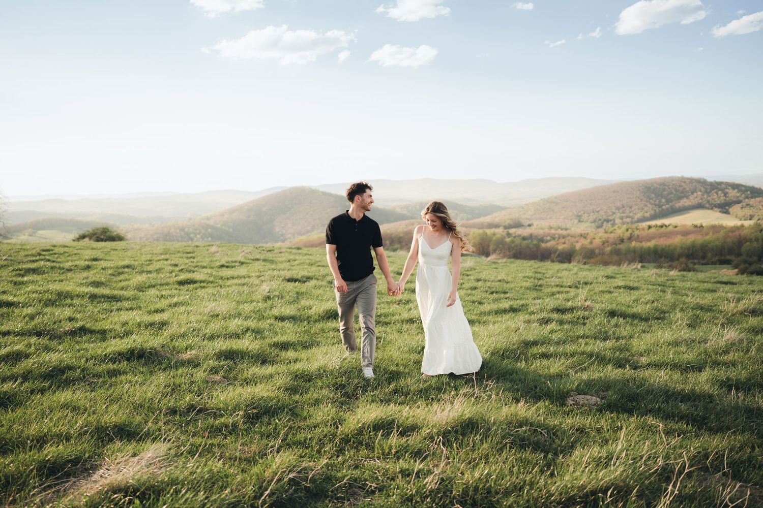 Couple holding hands walking through lush green hilltop meadow with scenic mountain valley views under blue sky.