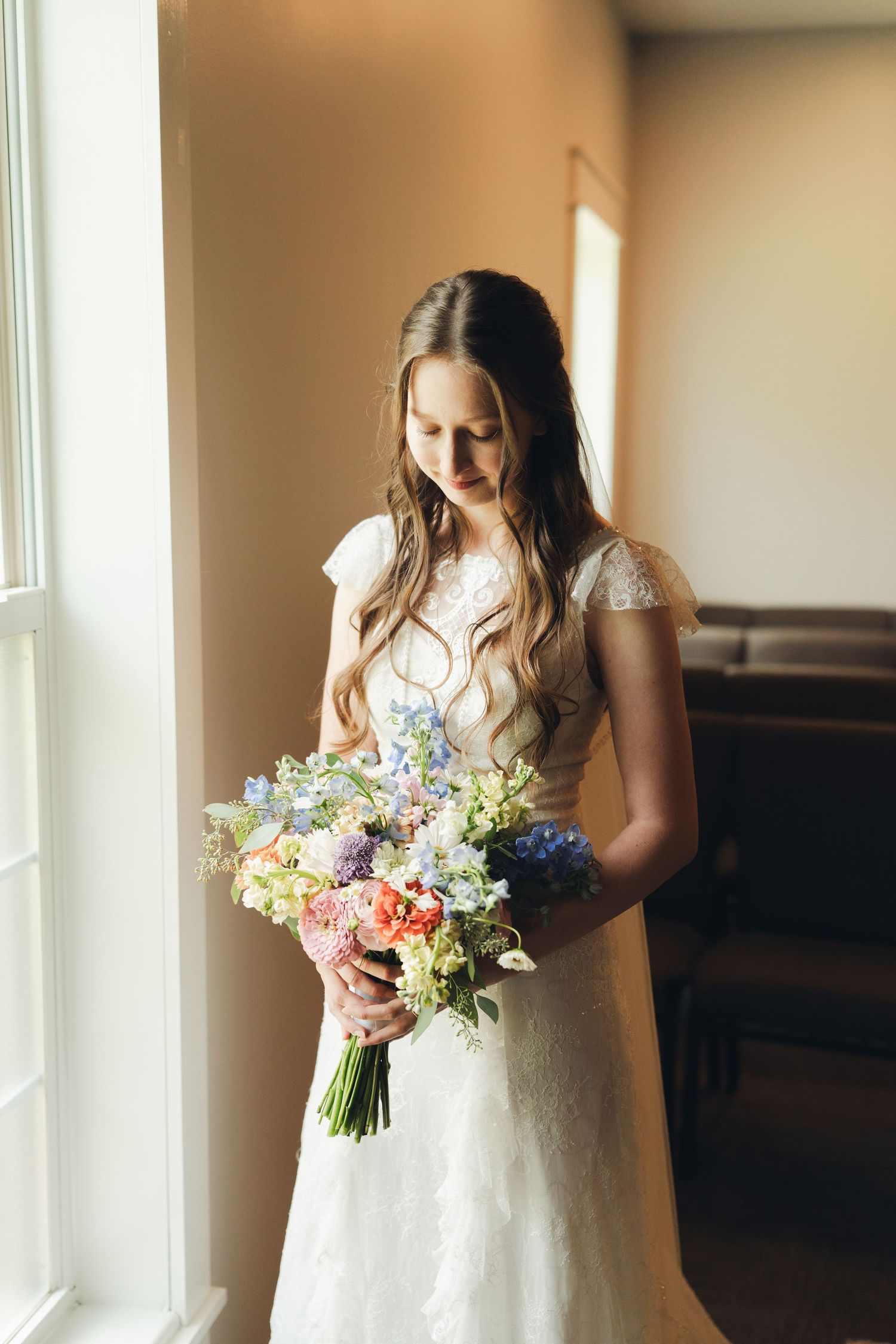 Bride in lace wedding dress holding colorful spring bouquet with blue, pink, and green flowers near a bright window.