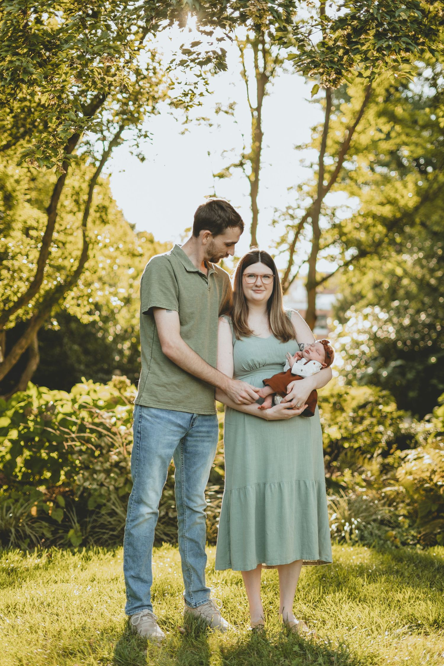 New parents stand together in sunny garden holding their newborn baby in warm golden light.
