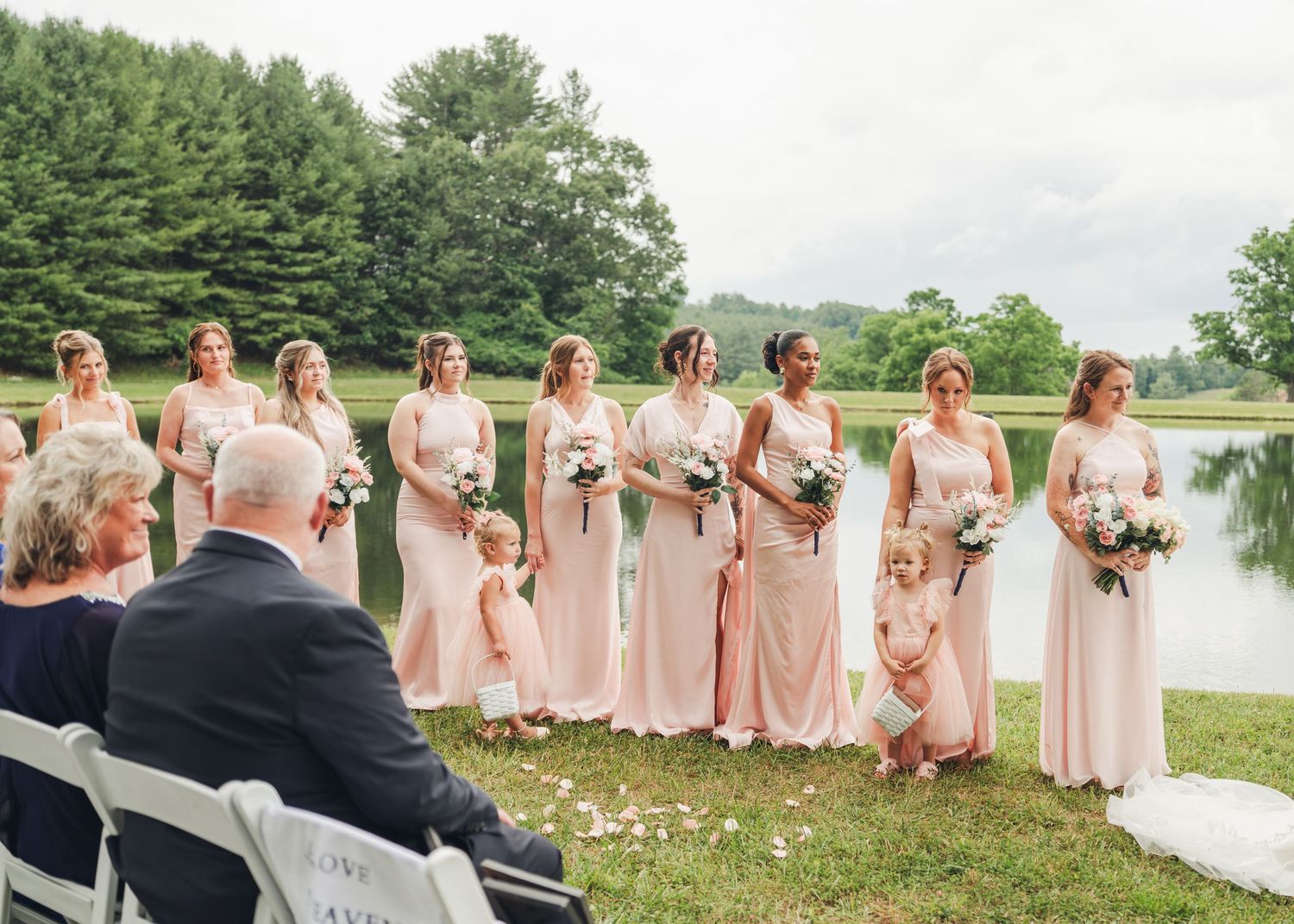 Bridesmaids in blush pink gowns holding bouquets stand outdoors near a pond during a wedding ceremony.