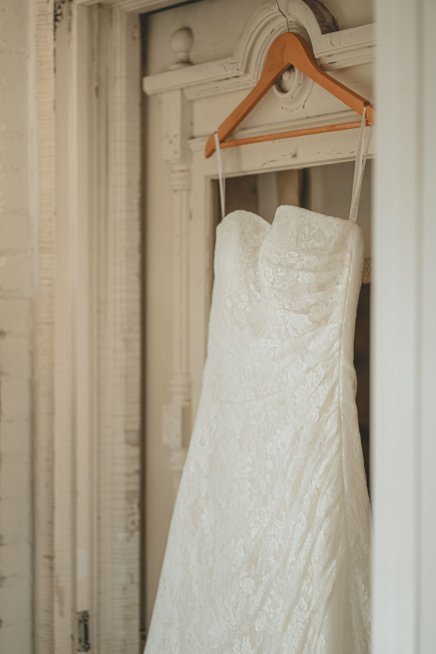 Strapless lace wedding dress hanging on wooden hanger against vintage white door frame.