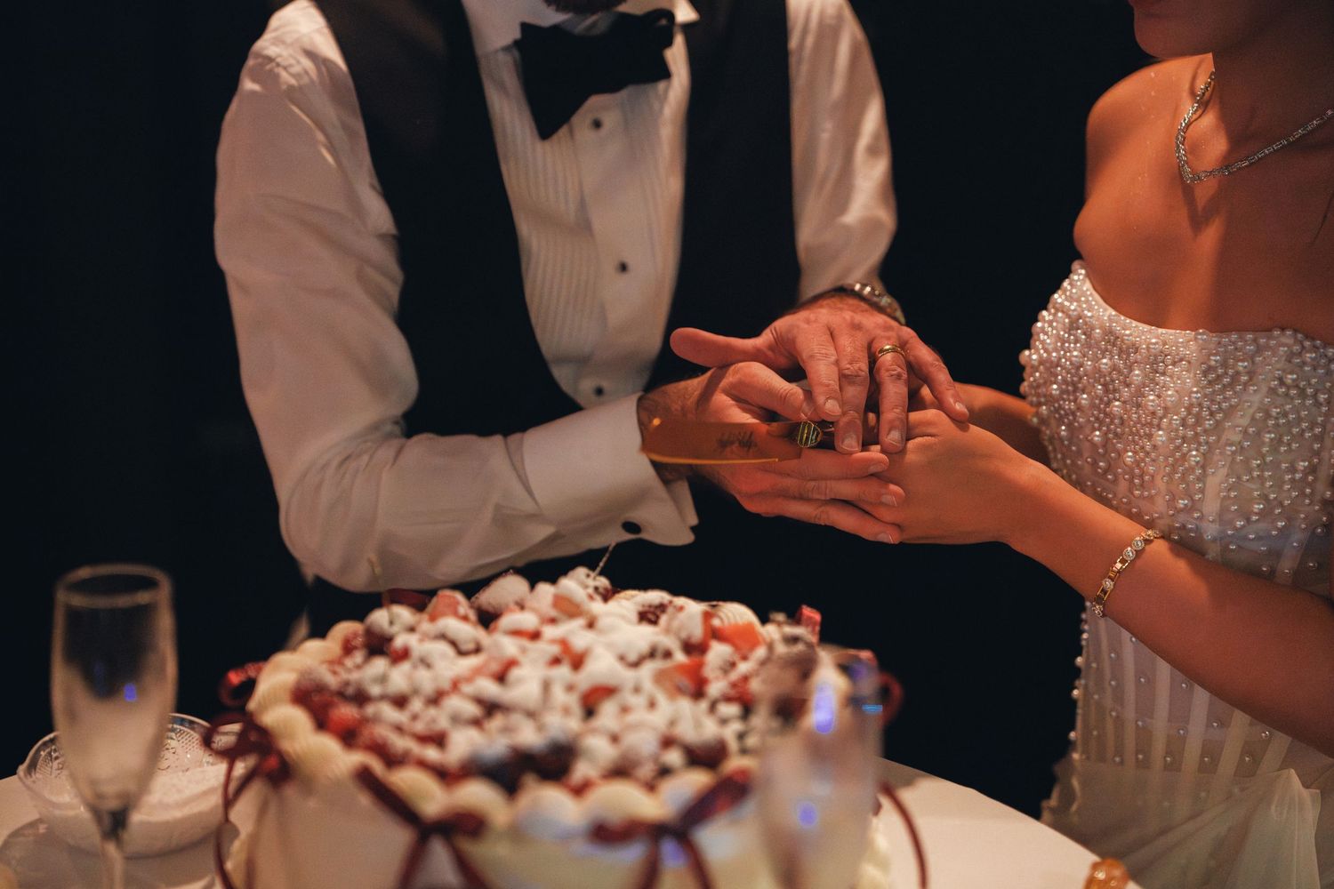 Bride and groom cut their wedding cake together, hands intertwined over a white cake with strawberry topping.