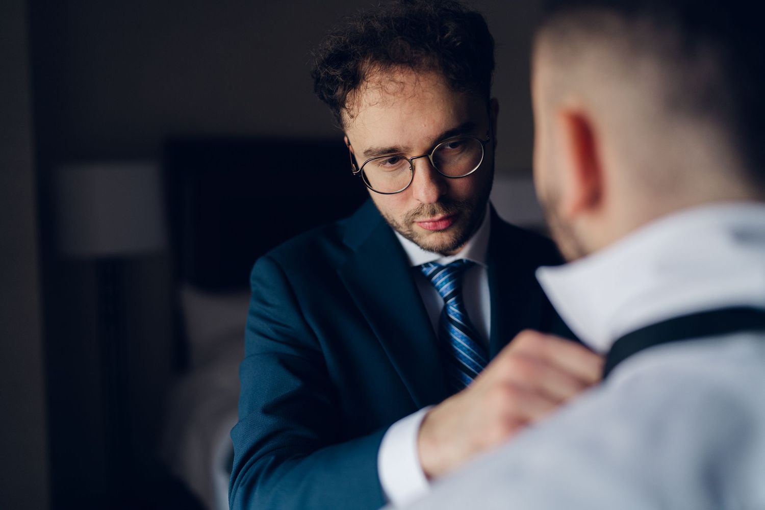 Groom in navy suit adjusting another mans suspenders during wedding day preparations.