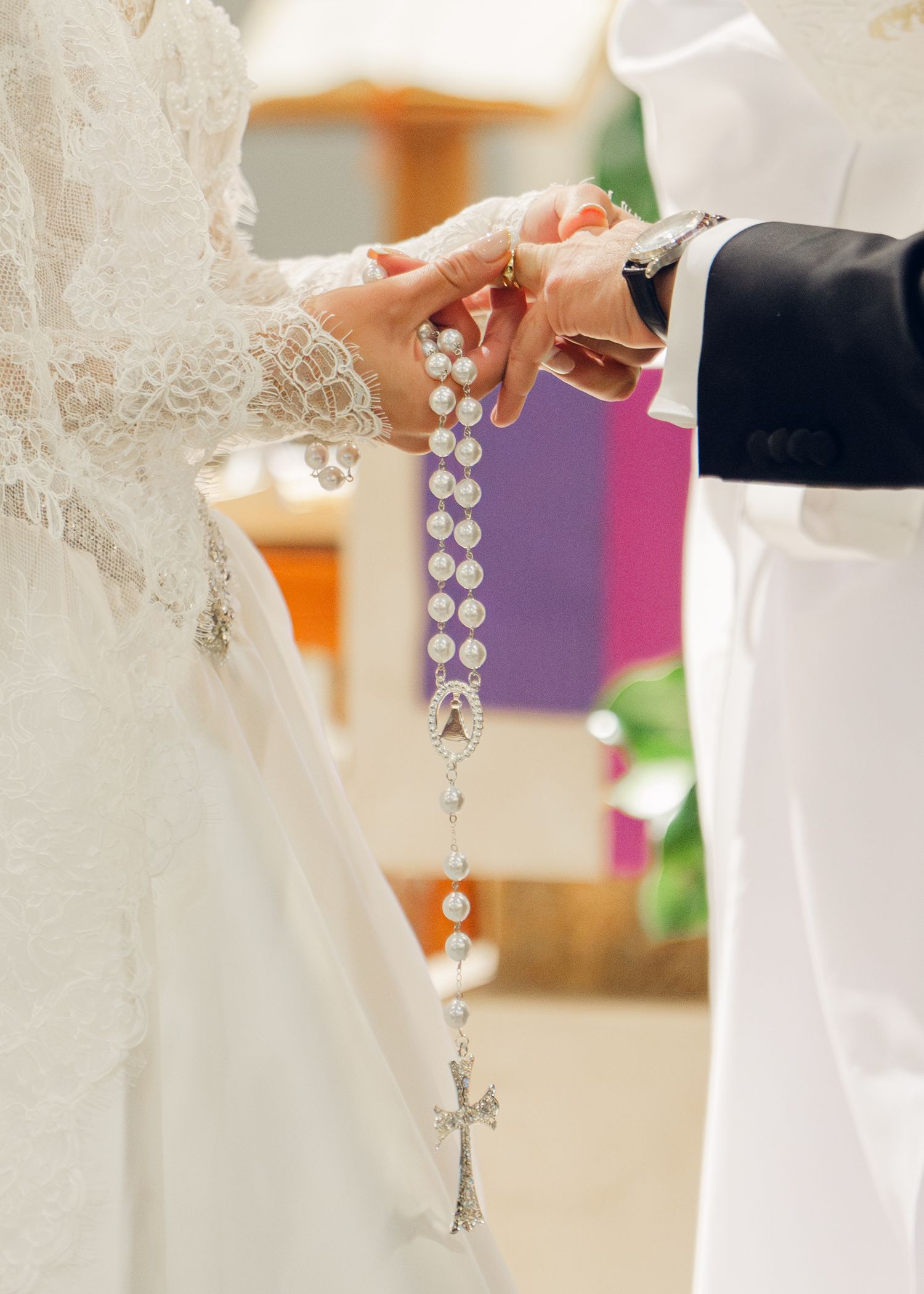 Close-up of bride and groom holding pearl rosary with cross during Catholic wedding.