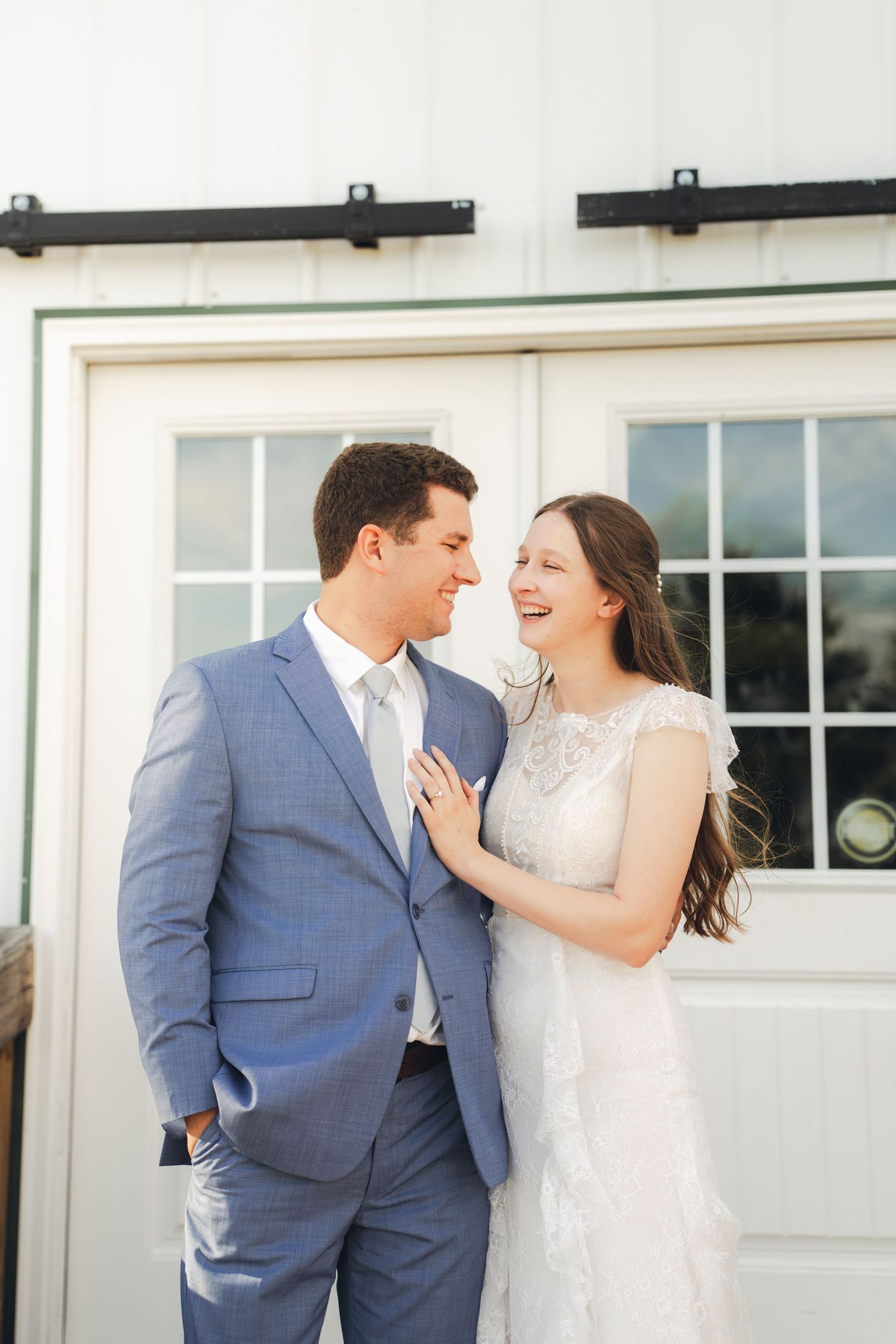 A smiling bride in a white lace dress and groom in a blue suit share a joyful moment outside a white barn.
