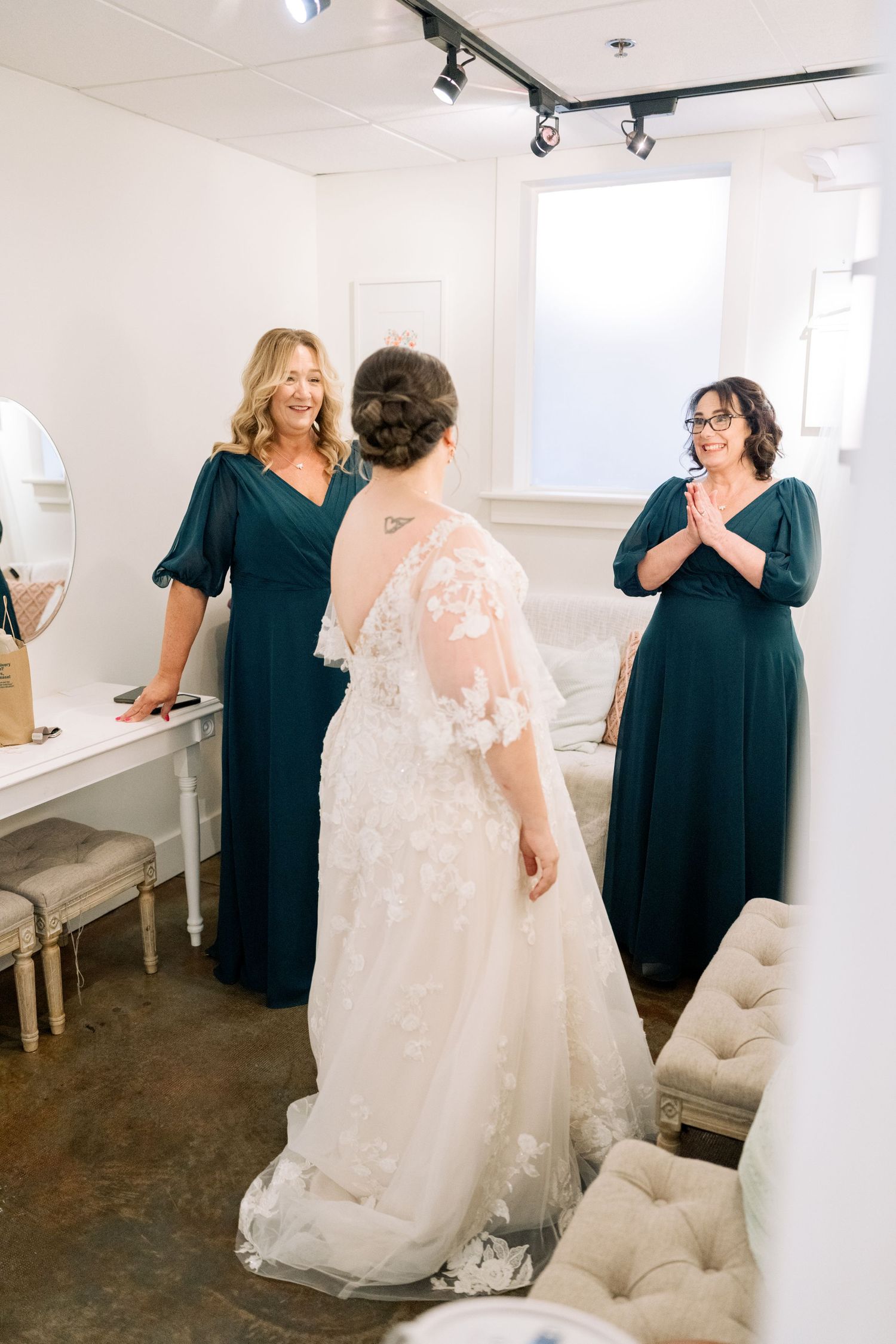 Bride in lace gown reveals dress to two bridesmaids in dark green dresses in bridal suite.