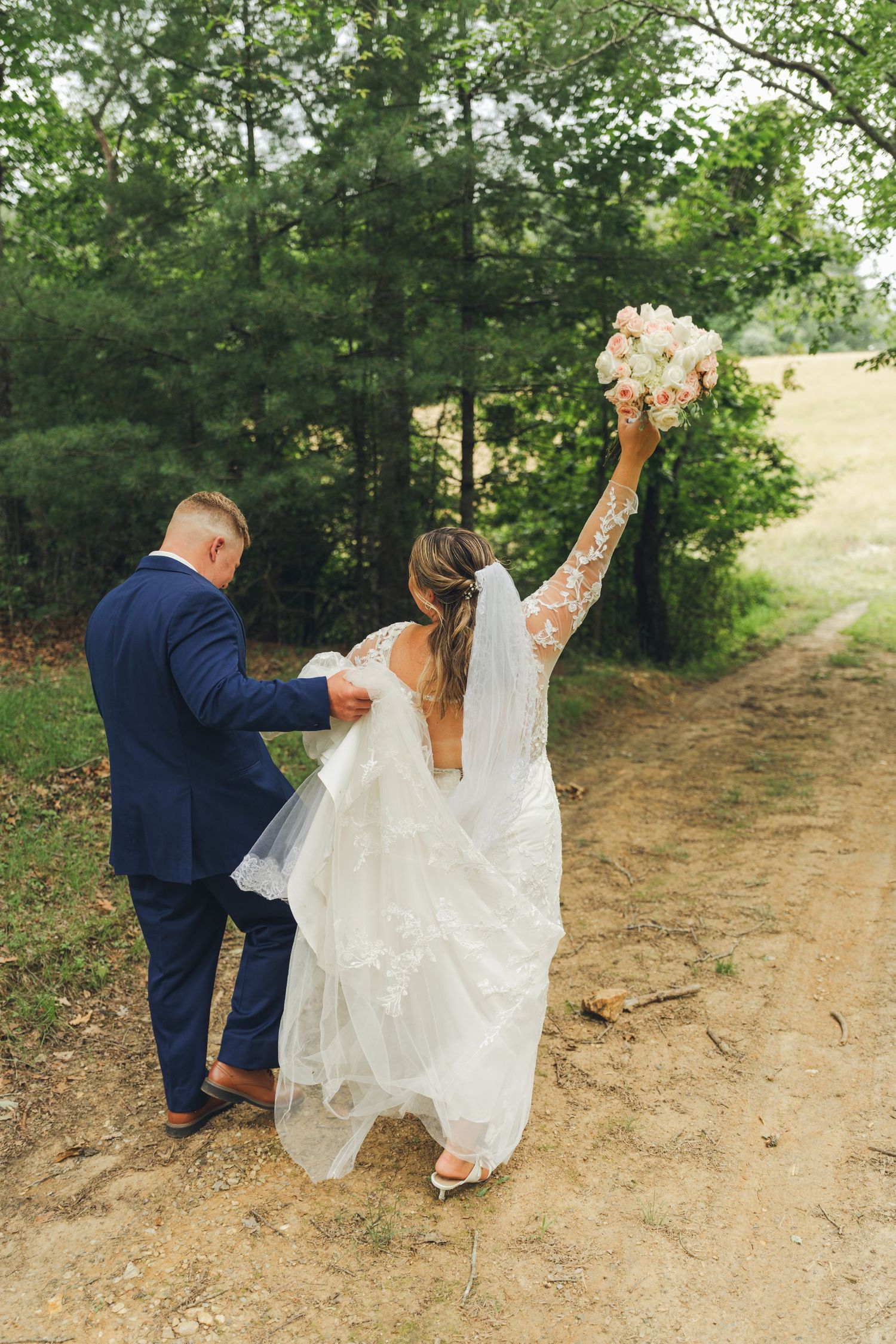 Bride raises bouquet of pink roses while groom holds her train on a rustic dirt path surrounded by lush green trees.