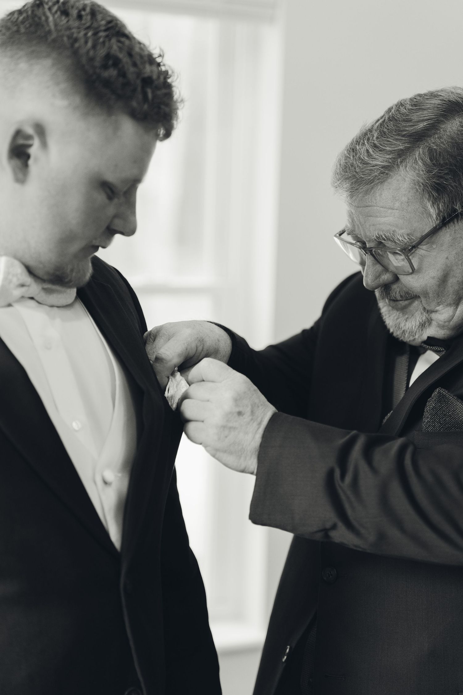 Black and white photo of older man pinning boutonniere on young groom's lapel before wedding ceremony.