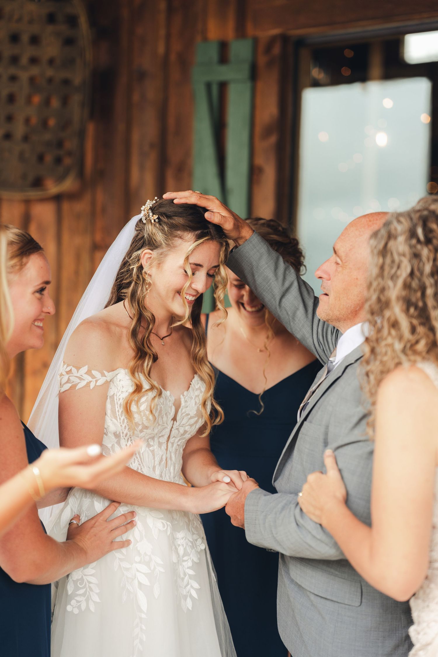 Bride in lace gown shares emotional first look moment with family inside a rustic barn venue.