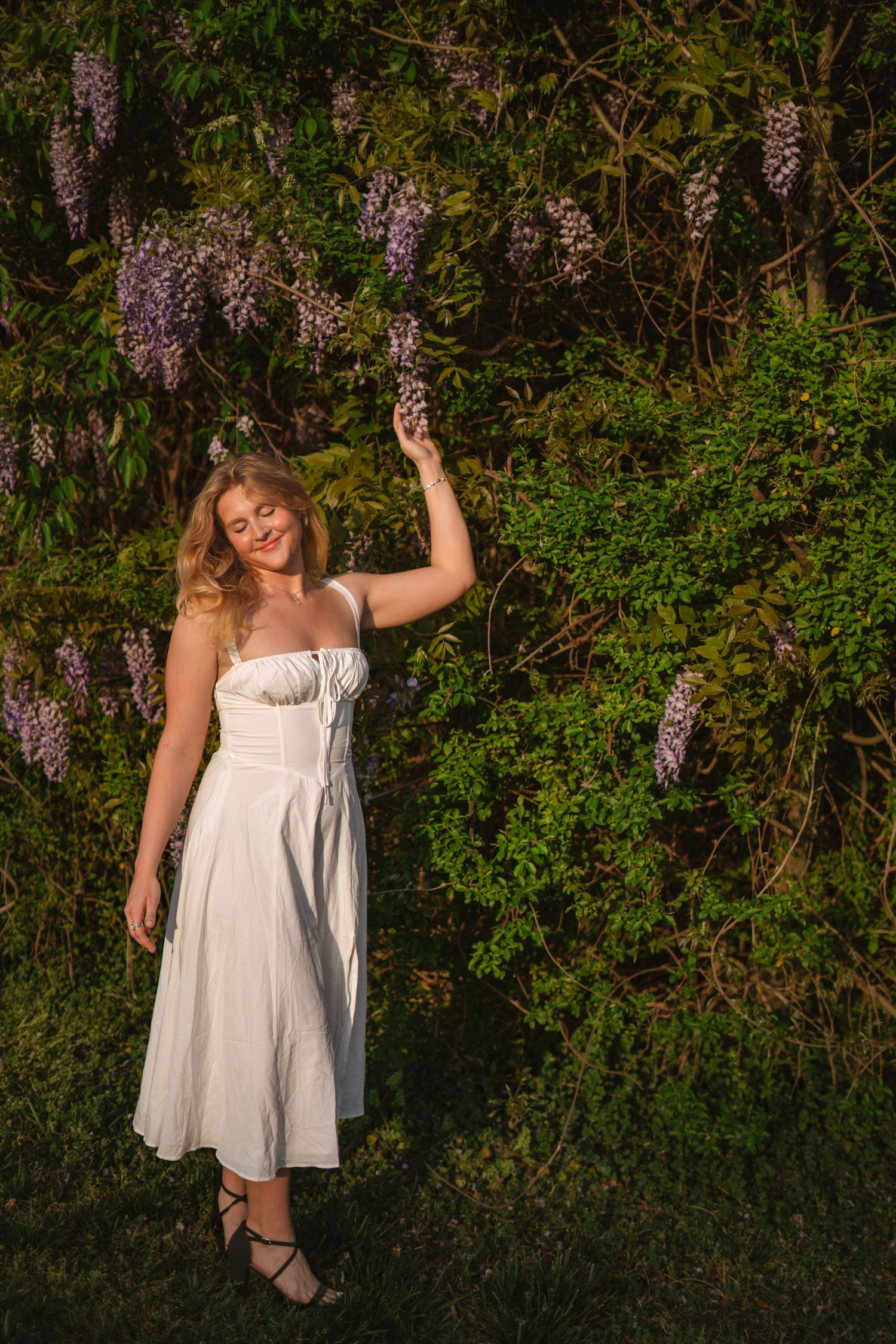 Young woman in white sundress reaching up to touch purple wisteria flowers in sunlit garden setting.