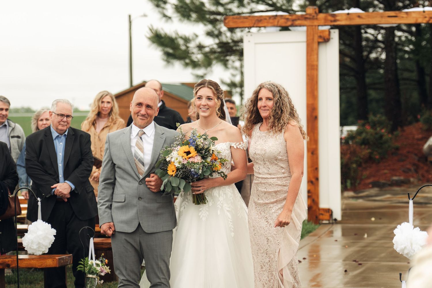 Bride walks down the aisle with parents at outdoor wedding ceremony, holding sunflower bouquet.