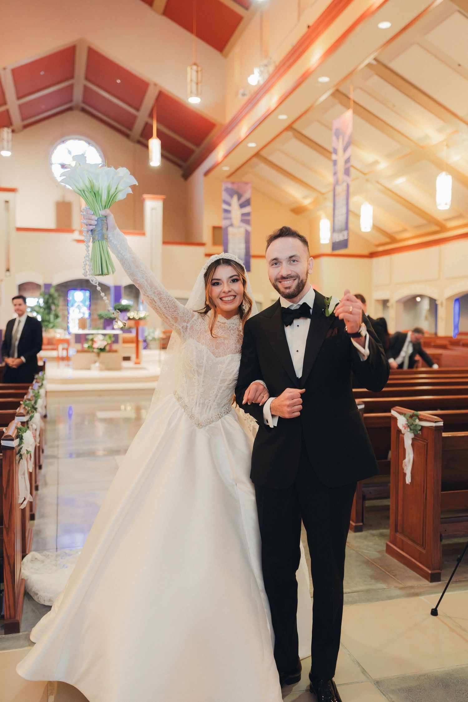 Joyful bride raises bouquet while walking down church aisle with groom after ceremony.