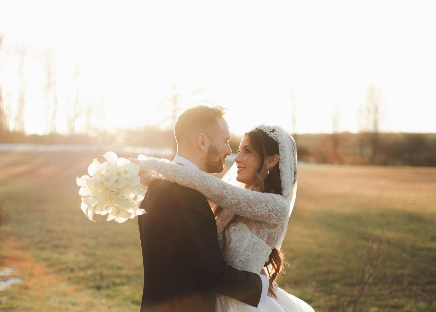 Groom lifting bride in a tender embrace at sunset, bride in lace gown holding white bouquet in open field.