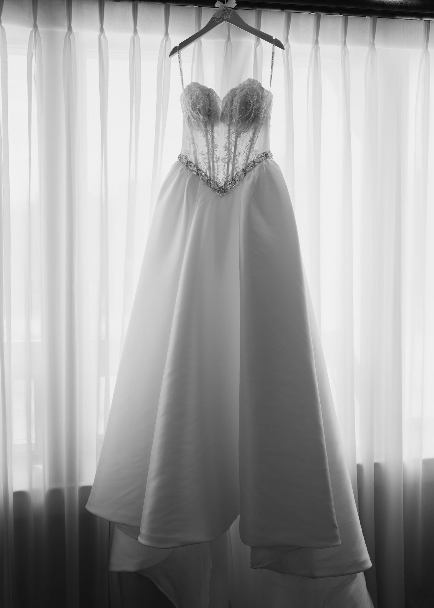 Black and white photo of a strapless beaded wedding gown hanging on a curtained window.