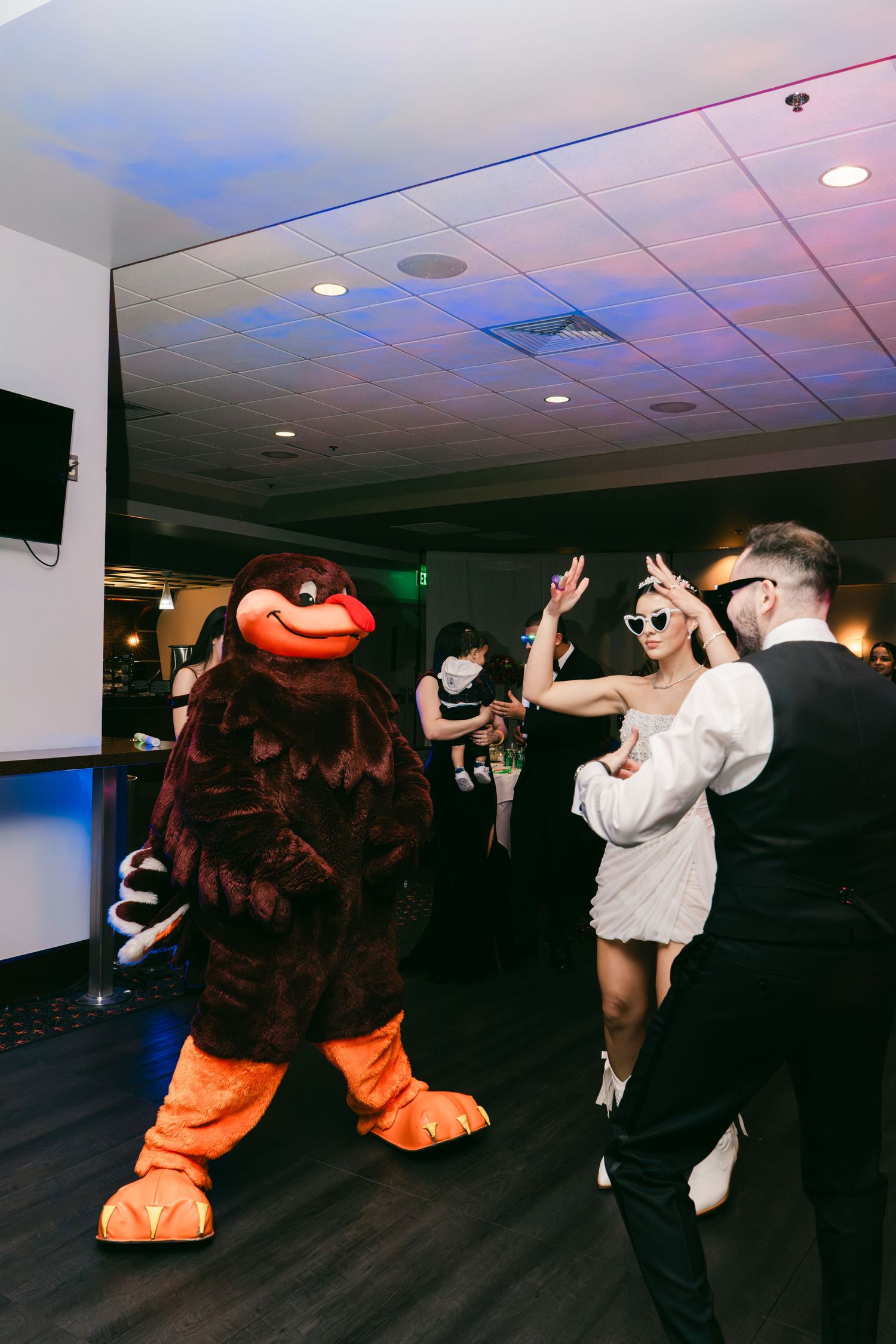Bride in short white dress dances with groom beside large brown bear mascot at reception.