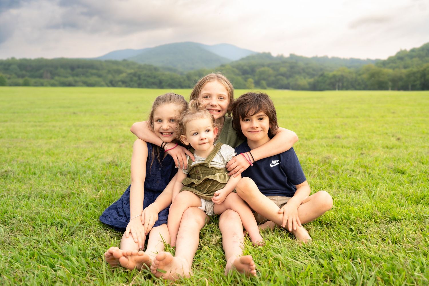 Four children sitting together on green grass with mountain backdrop, smiling for a family portrait outdoors.