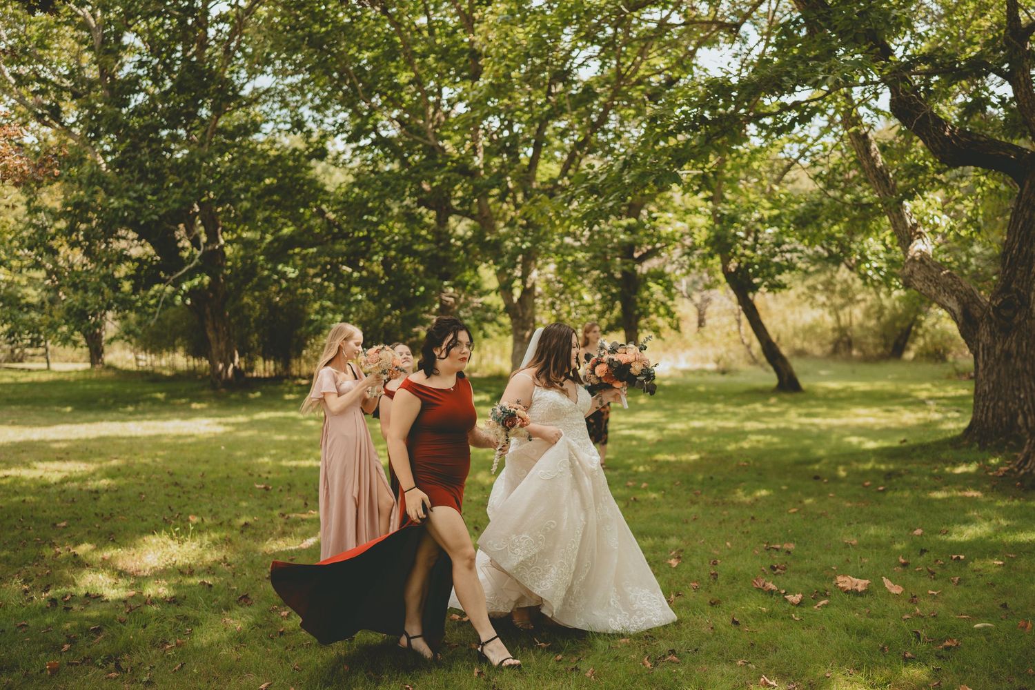 Bridesmaids in colorful dresses dance joyfully with bride in white gown in a sunny green park.