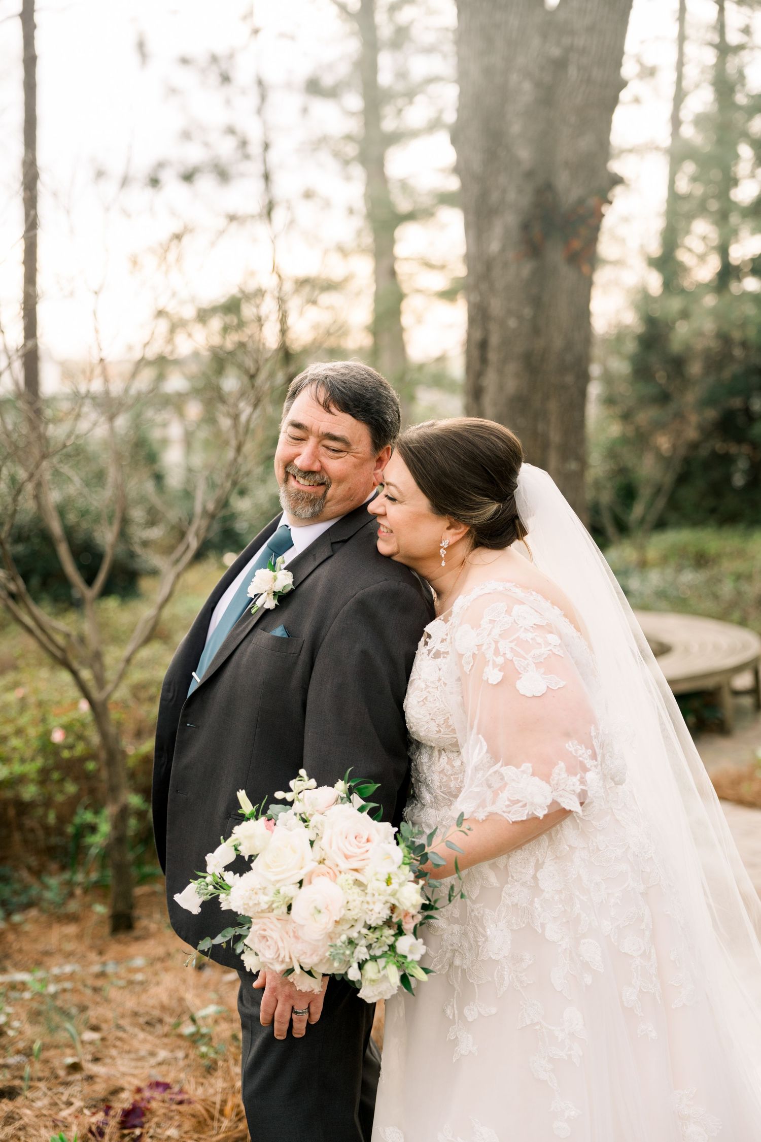 Bride resting her head on groom's shoulder as both smile joyfully in a wooded outdoor setting.