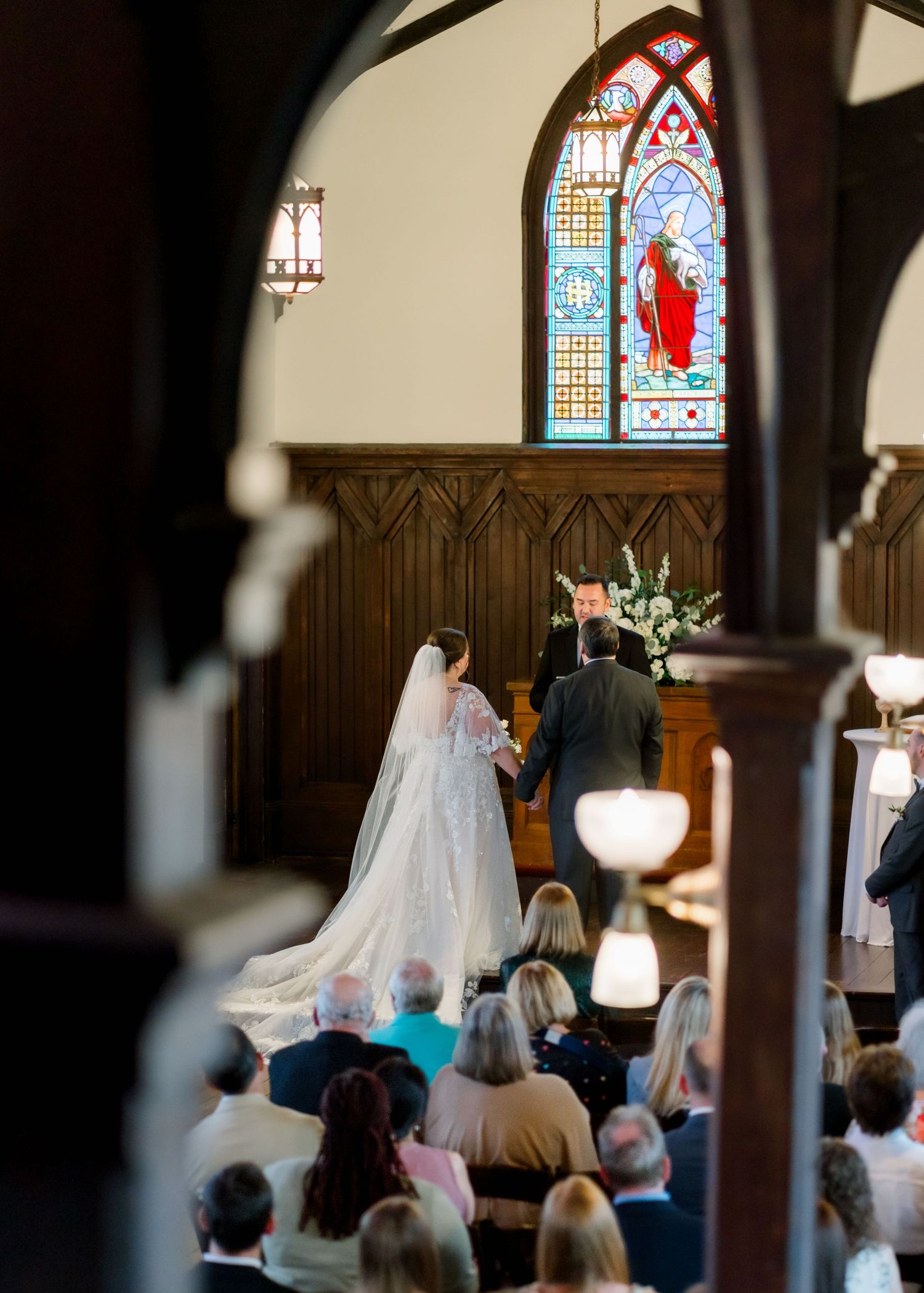 Bride and groom hold hands at altar framed by ornate stained glass window in elegant chapel.