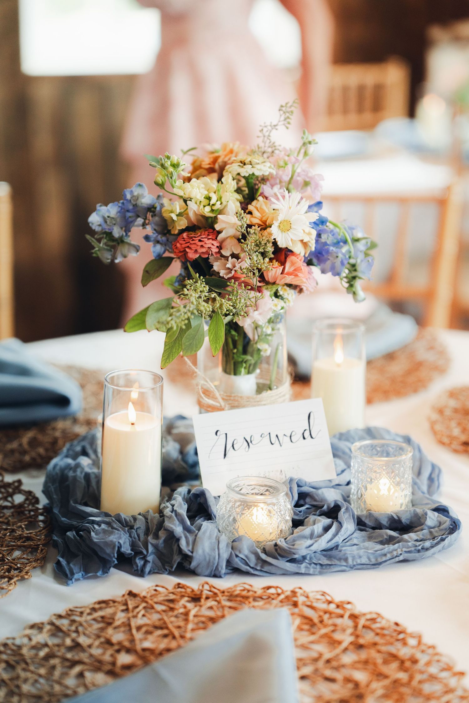 Rustic wedding reception table with floral centerpiece, white candles, and blue cheesecloth runner with reserved sign.