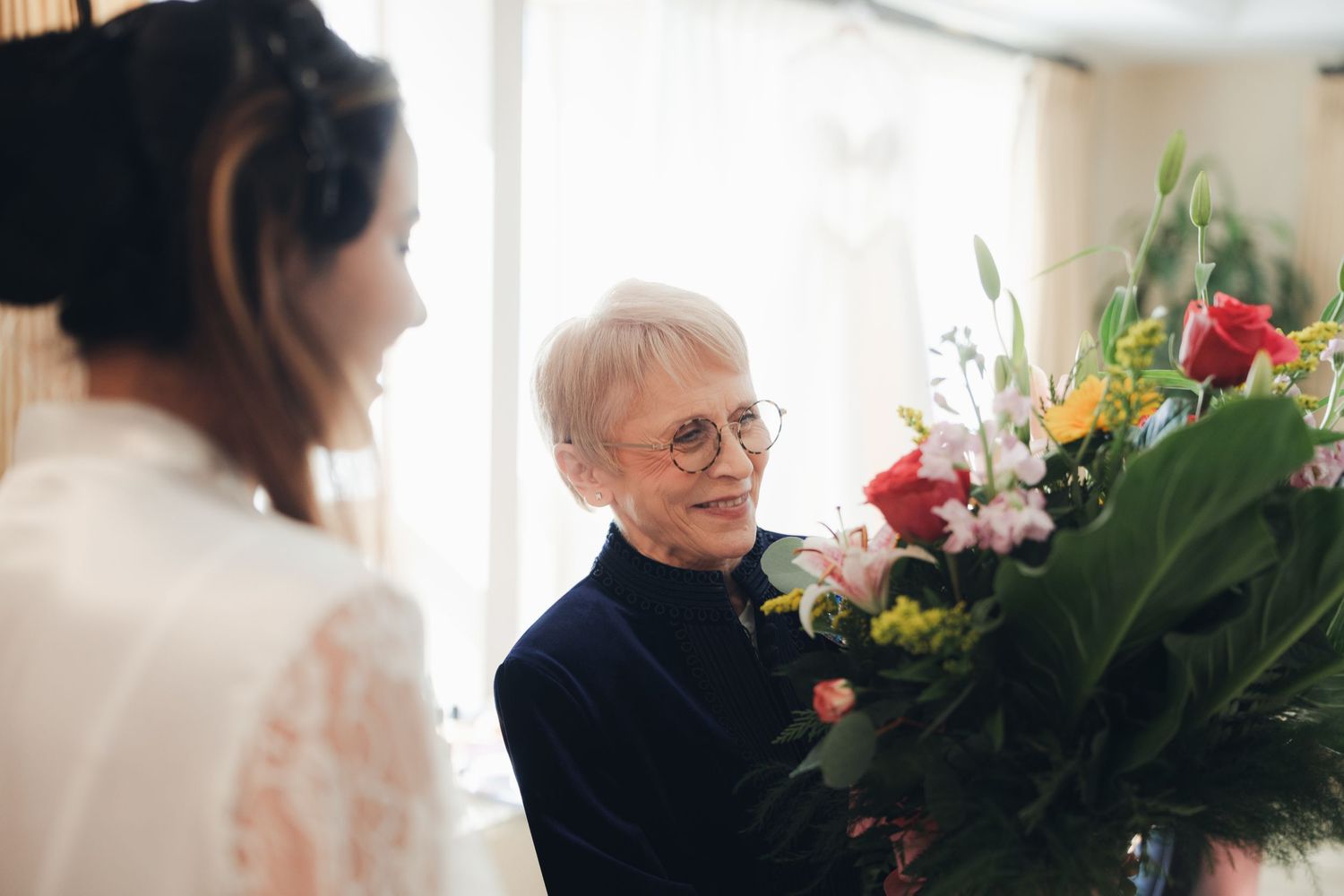 Elderly woman with glasses presenting a colorful flower bouquet to a bride in white.