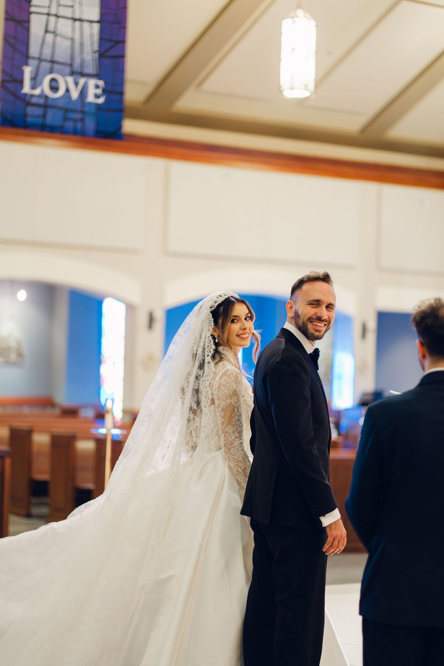 Smiling bride and groom turn toward camera inside church with LOVE banner overhead.