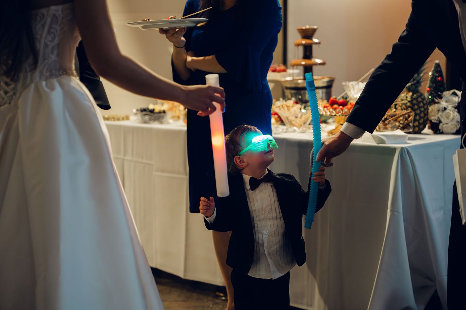 Young boy in tuxedo wearing light-up glasses holds glow stick at wedding reception party.