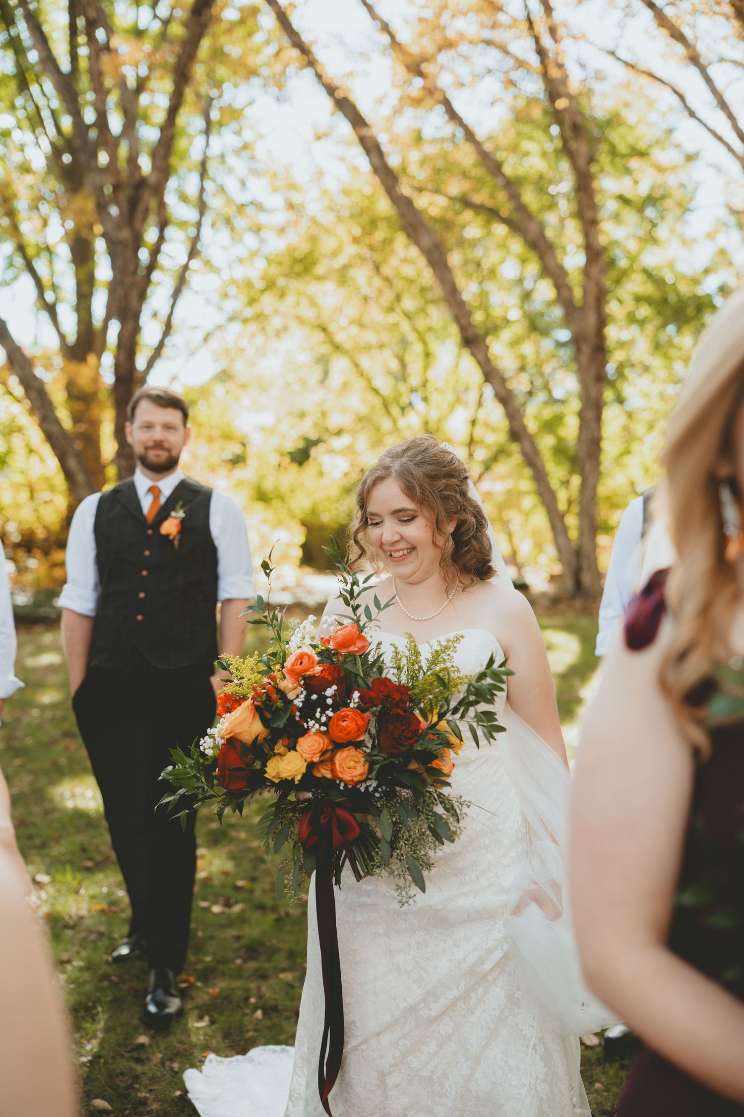 Smiling bride holding vibrant fall bouquet of orange and red flowers at outdoor wedding.