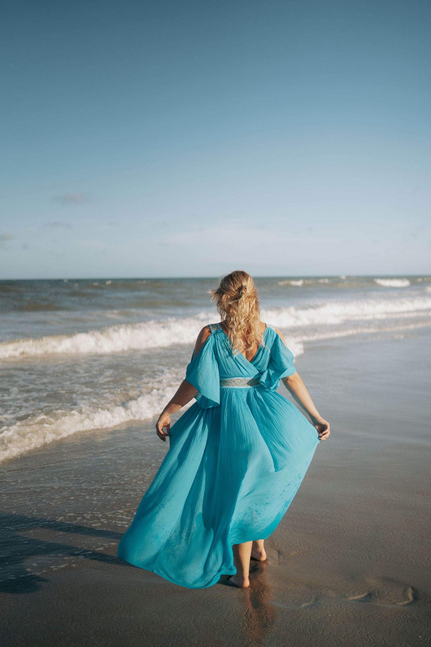 Woman in flowing turquoise dress walking barefoot along ocean shoreline on sunny day.