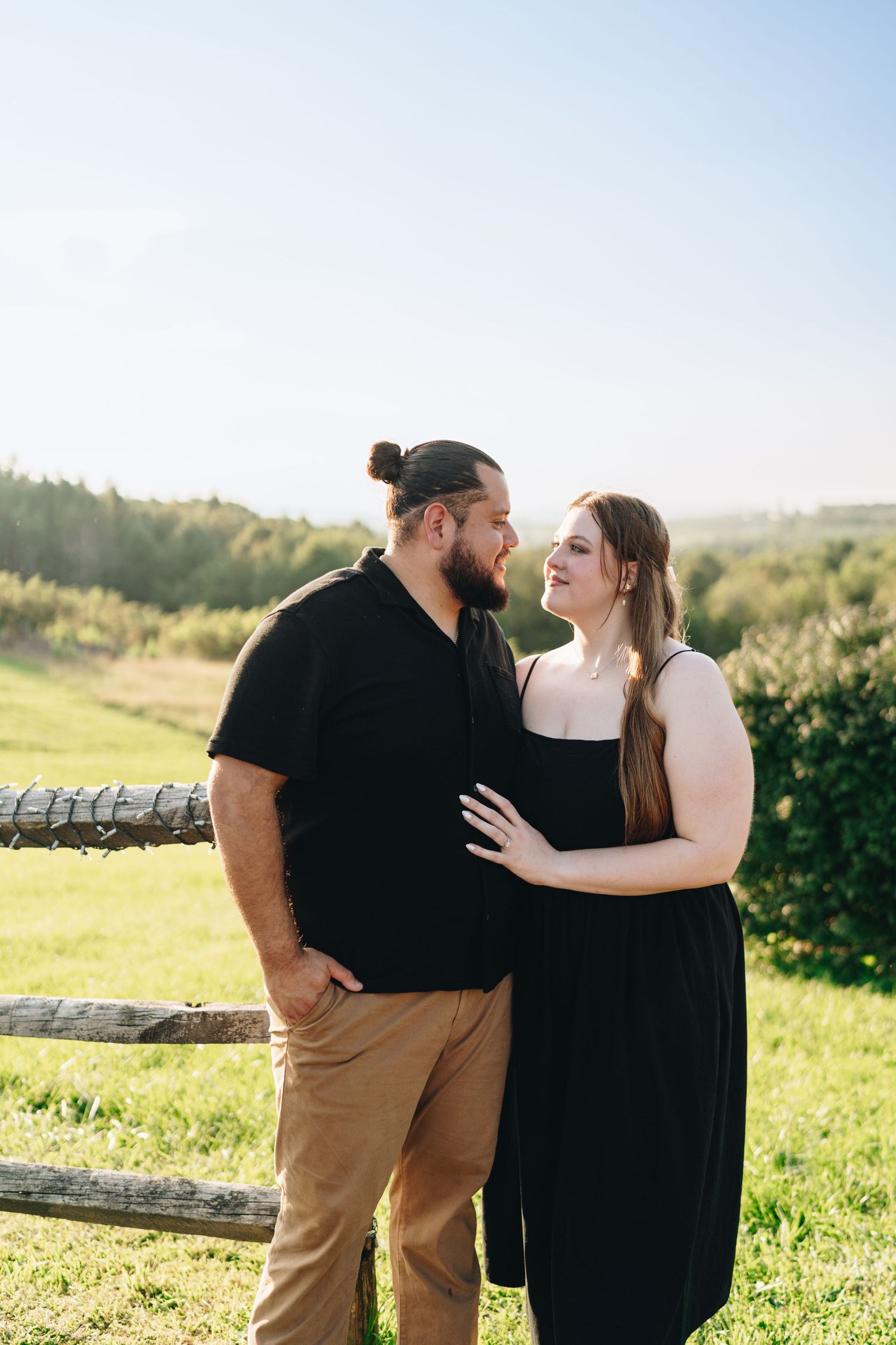 Smiling couple in black outfits gazing at each other near a wooden fence on a sunny day.