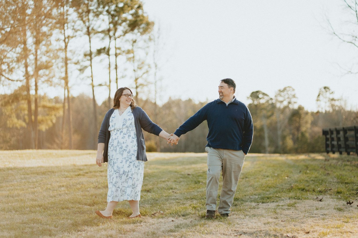 Couple walking hand-in-hand through a sunlit field, smiling at each other near pine trees.