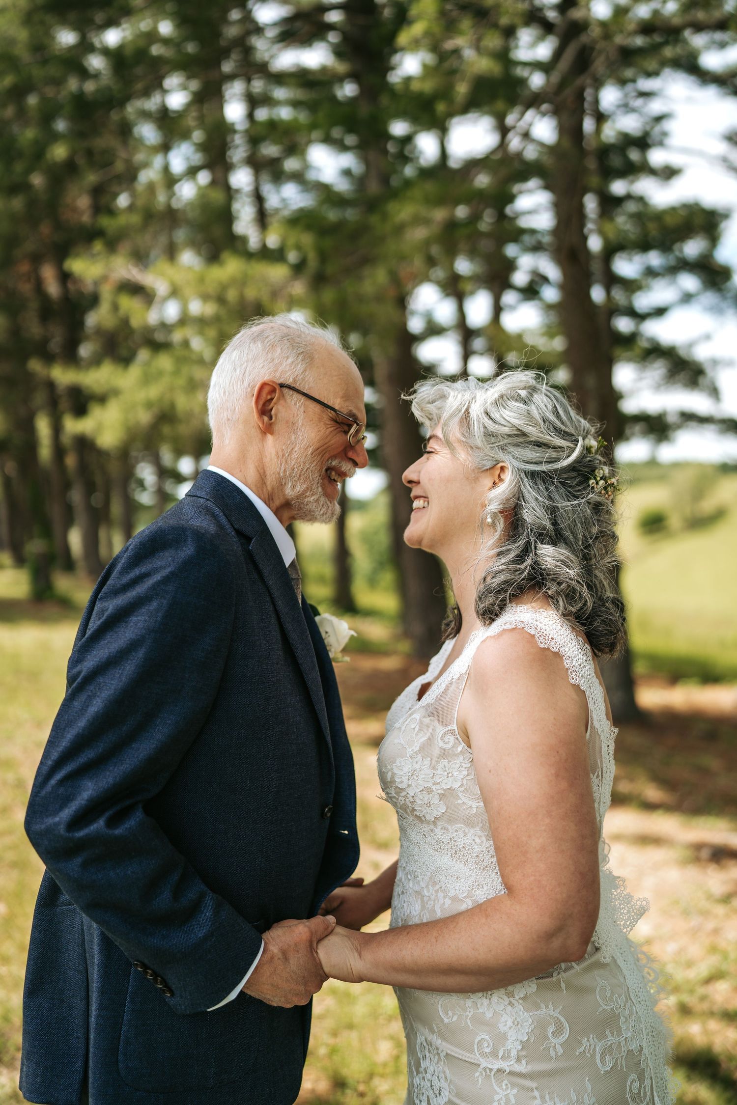 Older bride and groom holding hands and smiling at each other outdoors among tall pine trees.