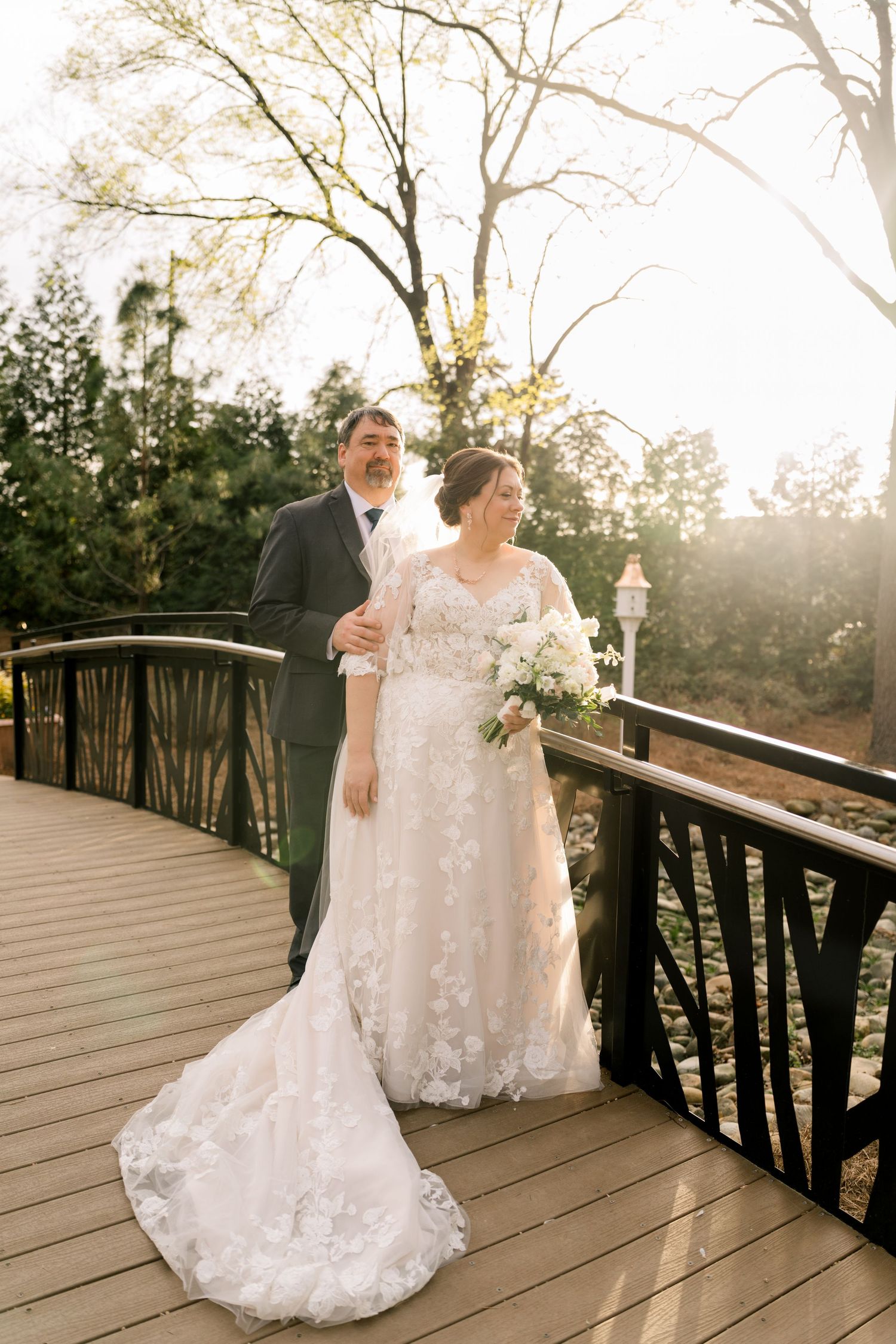 Bride and groom standing together on a wooden bridge, bathed in warm golden sunset light outdoors.