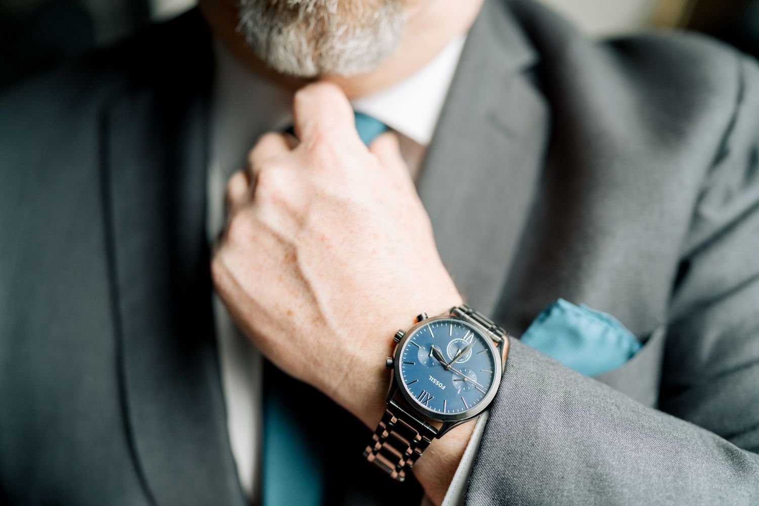 Close-up of groom adjusting teal tie wearing elegant blue-faced watch and gray suit.