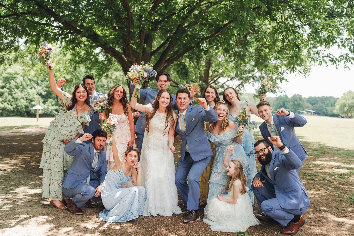 Wedding party group photo under large tree with groomsmen in blue suits and bridesmaids in floral dresses.