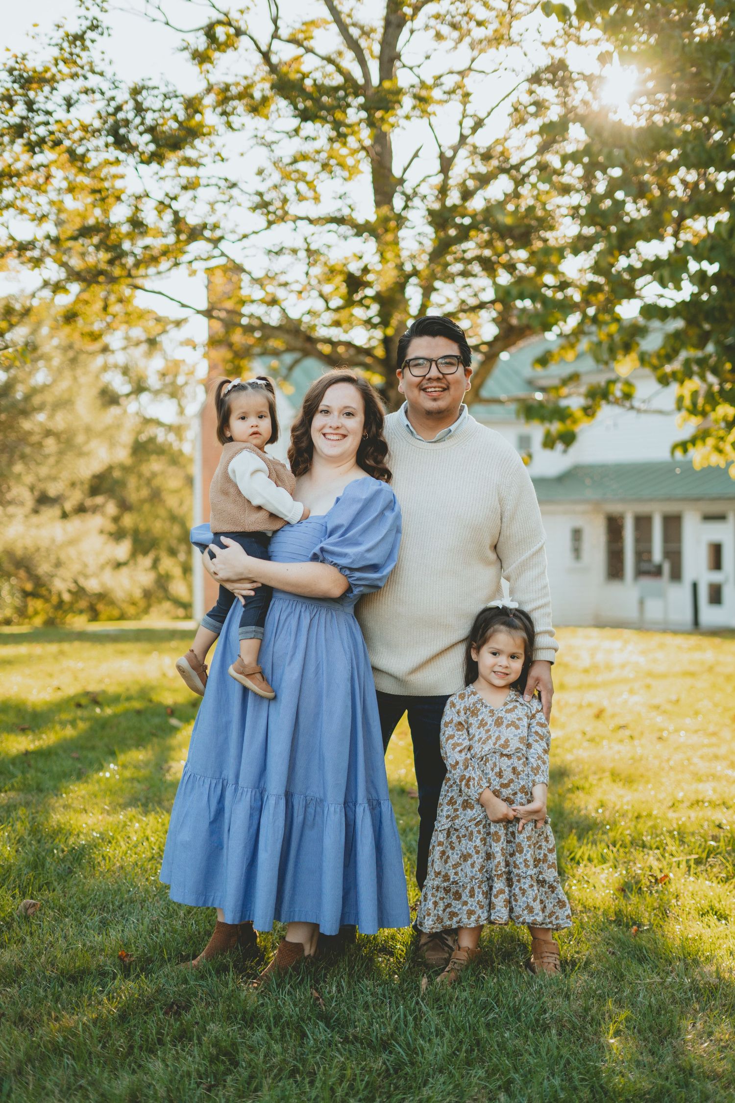 Happy family of four posing in golden sunlight on green lawn with white farmhouse backdrop.