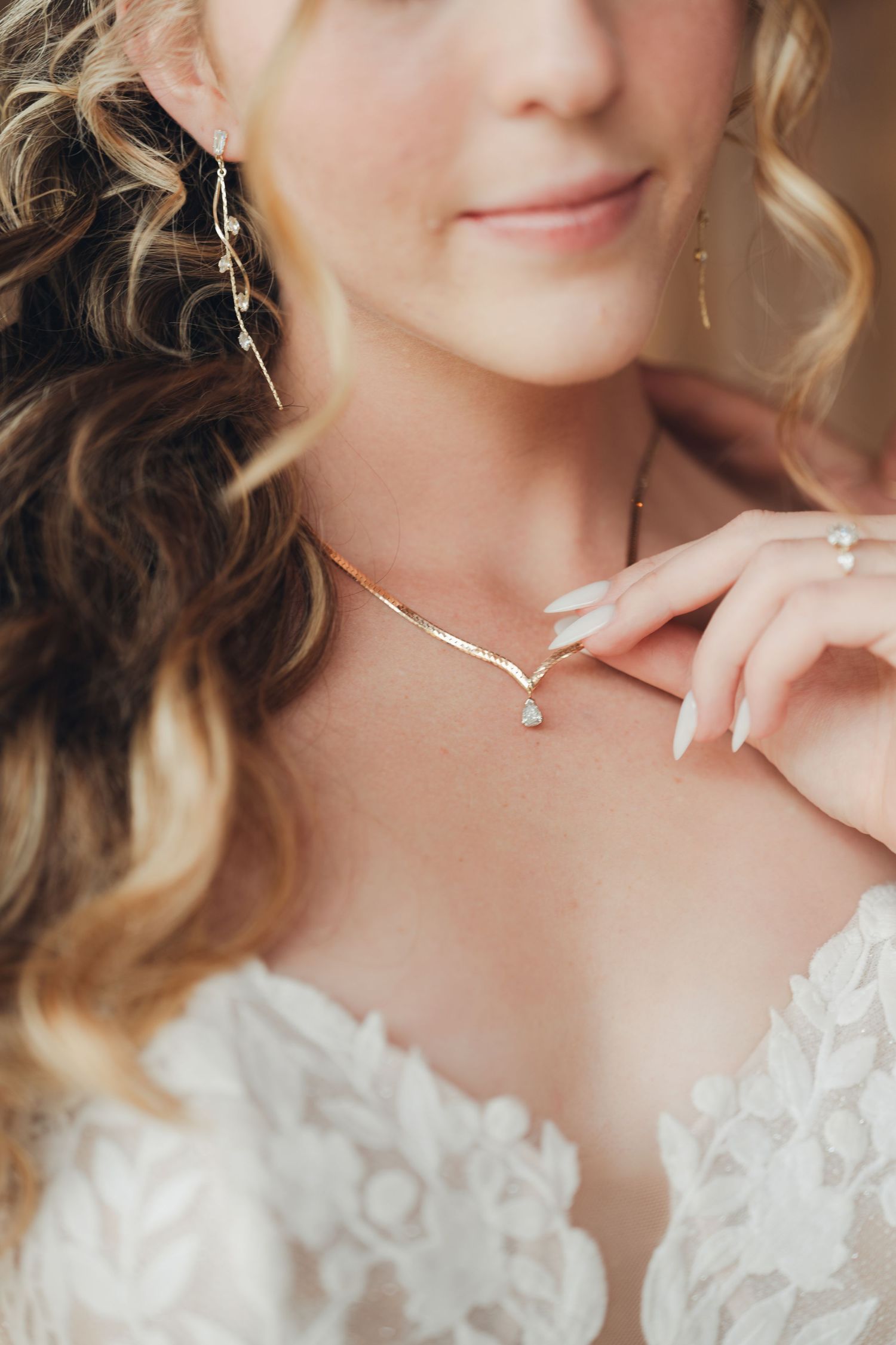 Bride wearing elegant gold V-shaped necklace with pearl drop, lace wedding dress, and dangling earrings, touching jewelry softly.