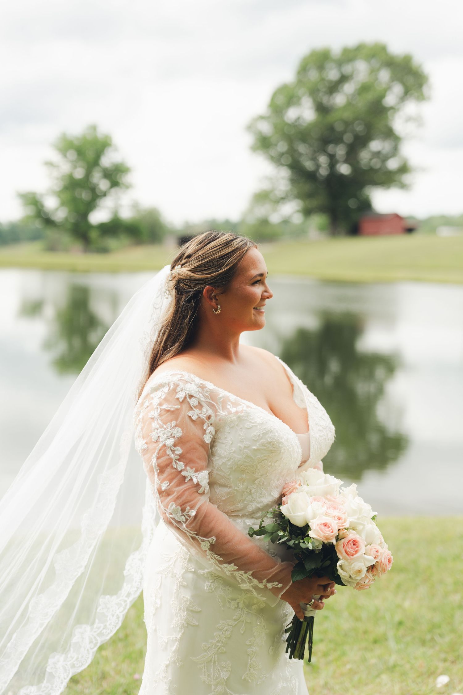 Bride in lace gown and veil holds pink bouquet, smiling by a serene pond with green trees reflected in water.