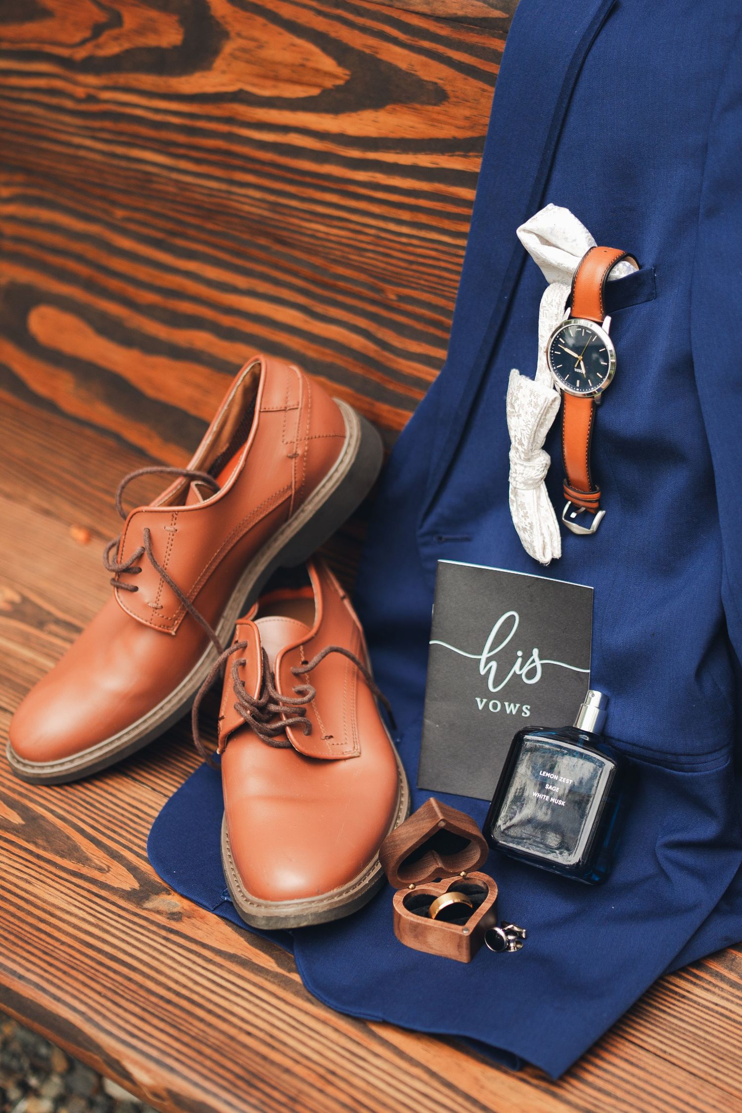 Groom's wedding accessories flat lay with tan leather shoes, blue suit, watch, cologne, and His Vows book on wood floor.