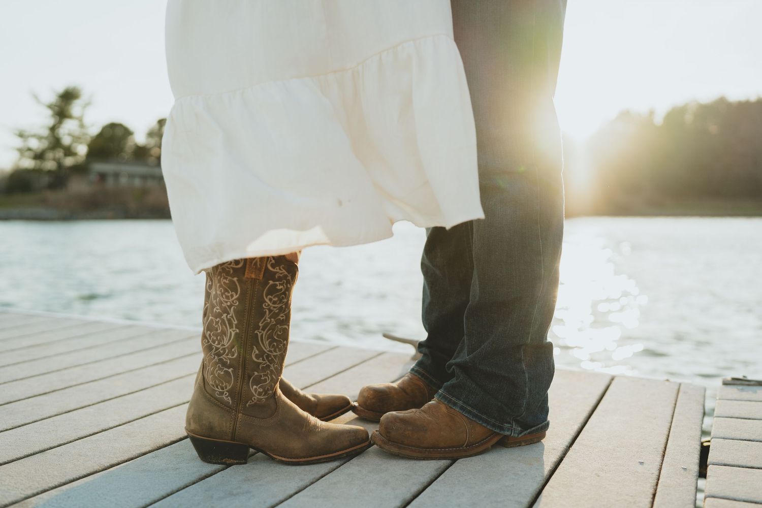 Close-up of cowboy boots and dress shoes on a wooden dock during a lakeside engagement session.