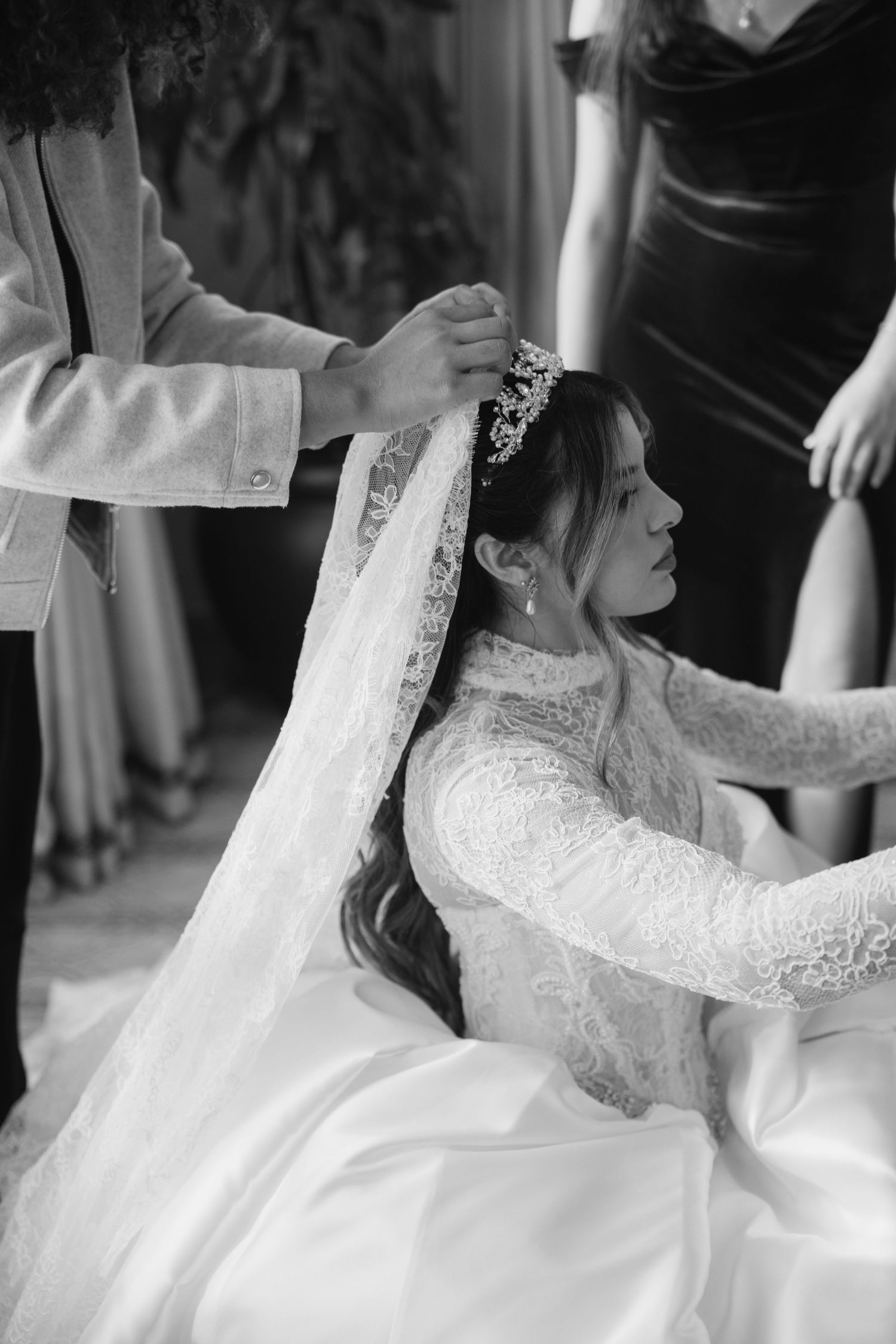 Black and white photo of bride having lace veil placed on tiara by attendants.